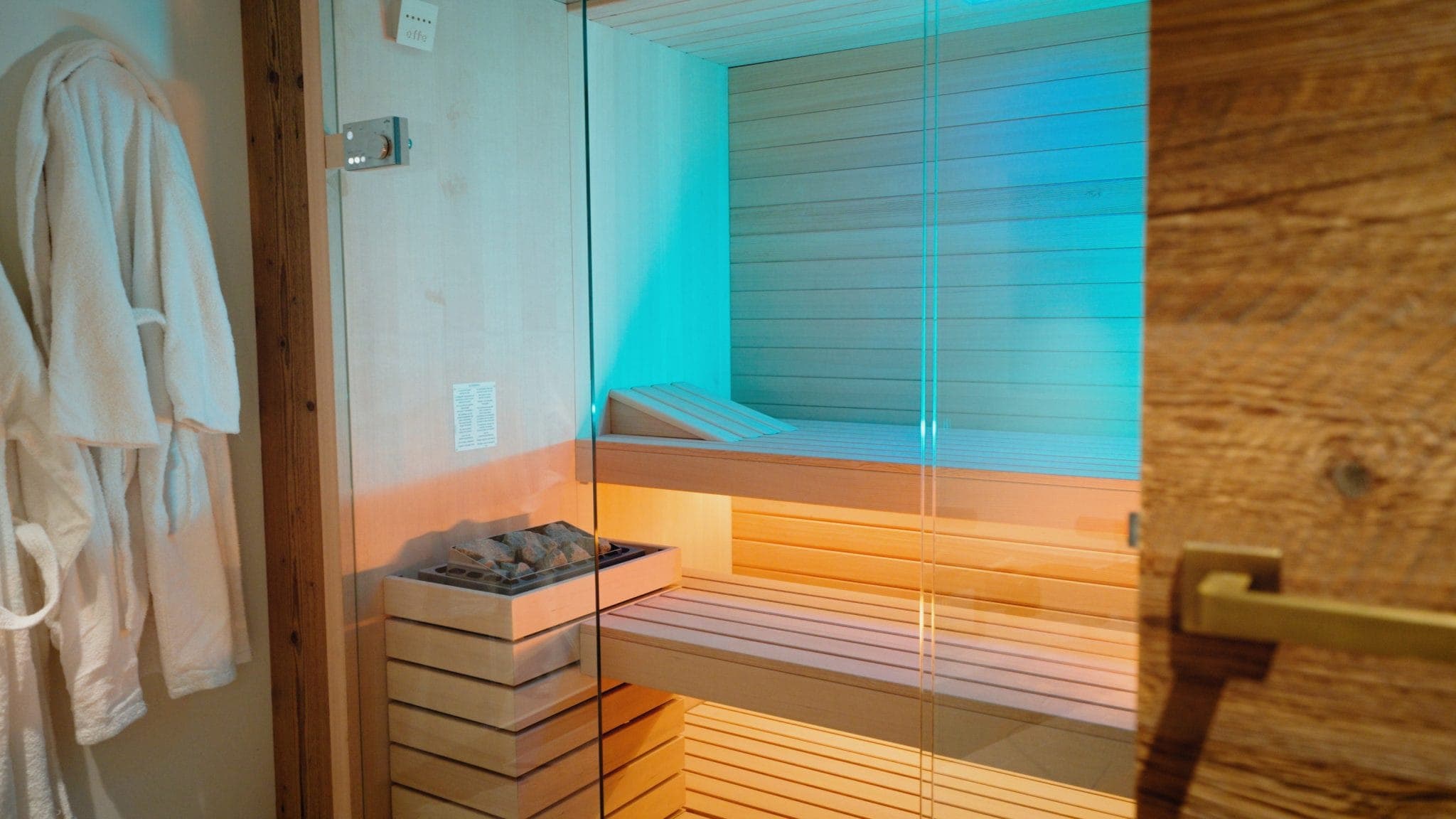 A cozy wooden sauna with a glass door, warmly lit from inside. Soft ambient lighting creates a relaxing atmosphere. A stack of stones is ready for use, and two plush white robes hang nearby.