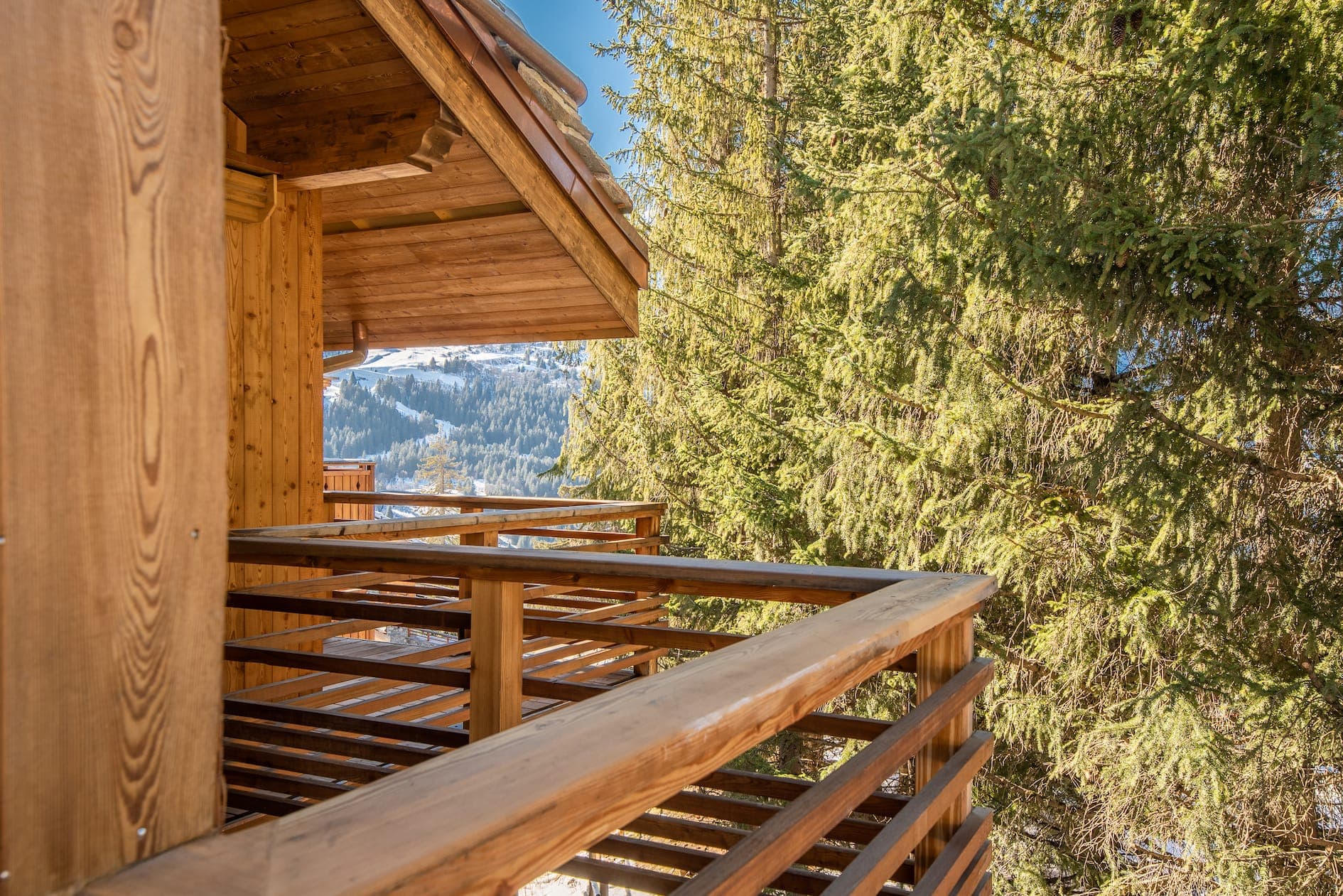 Wooden chalet balcony overlooking a snowy mountain landscape with tall pine trees nearby. The sky is clear and blue, enhancing the serene outdoor winter atmosphere. The structure features warm wood tones and rustic design elements.