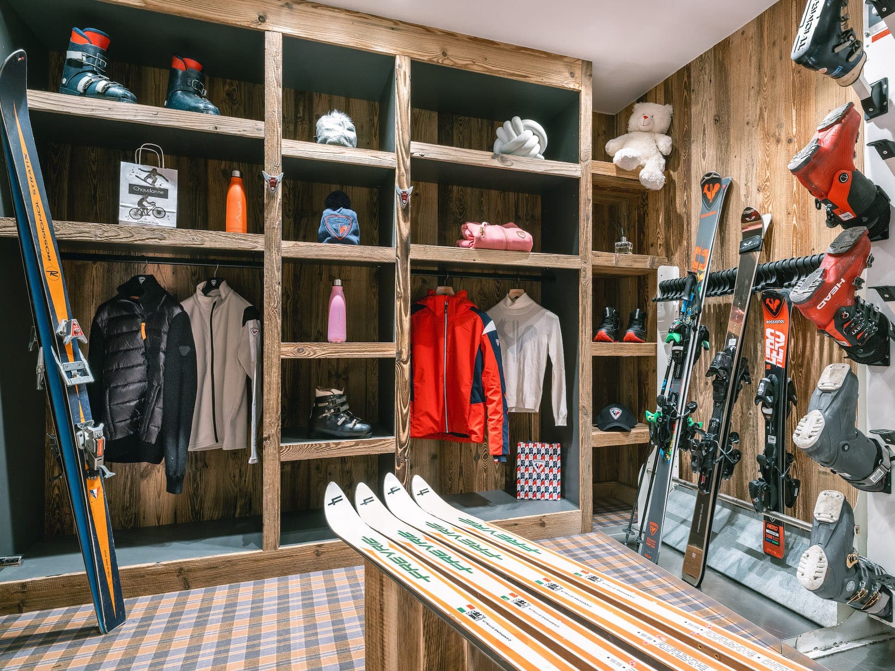 A comfortable ski room with wooden shelves displaying jackets, boots, helmets and accessories. Several skis are lined up in the foreground. Other skis and poles are displayed on the walls. A teddy bear and colorful water bottles add a playful touch.