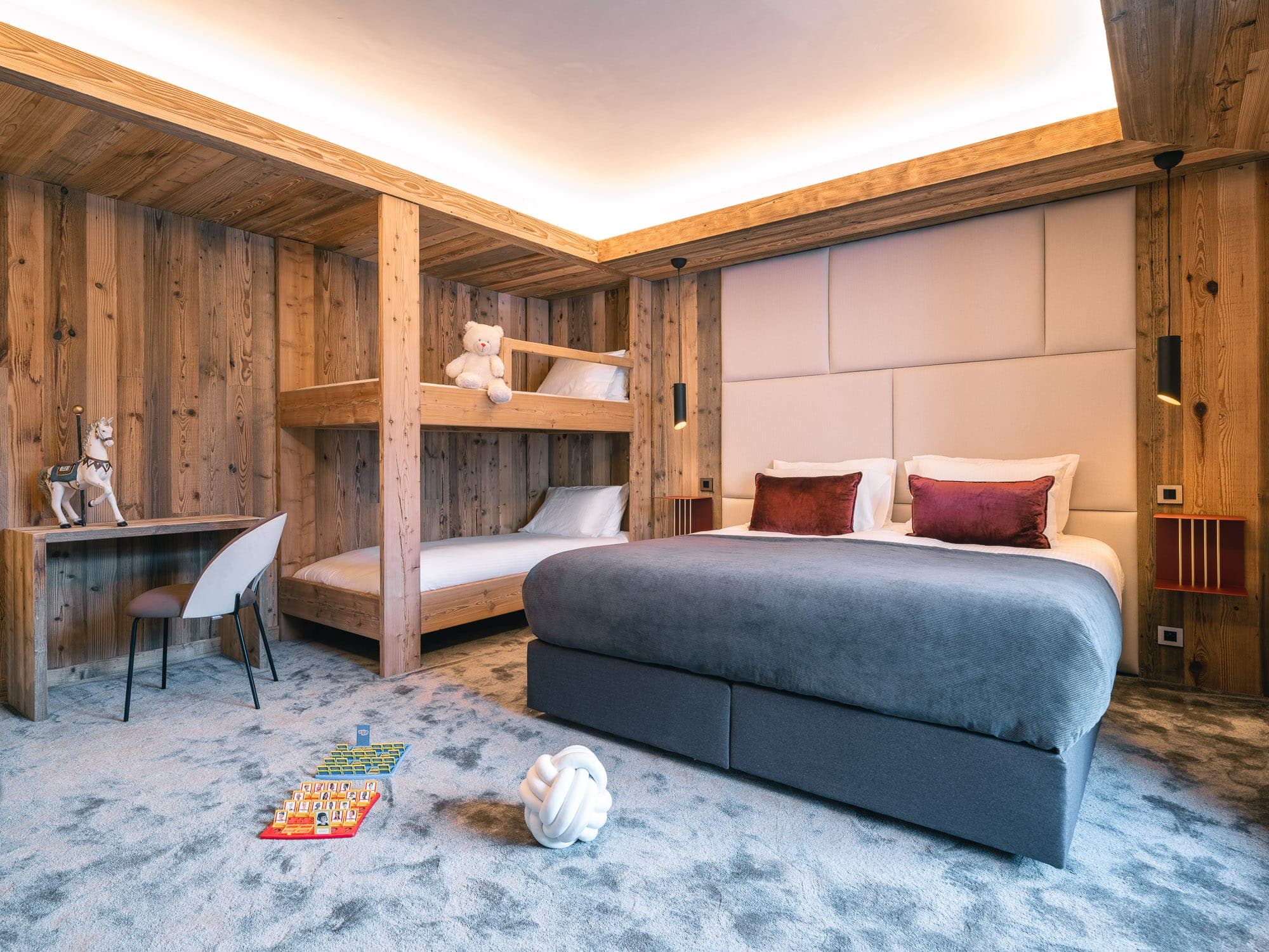 Cosy room with wooden walls, a large bed with gray and white bedding, bunk beds with teddy bears, a small desk and chair, and decorative objects on the floor. Soft lighting creates a warm atmosphere.