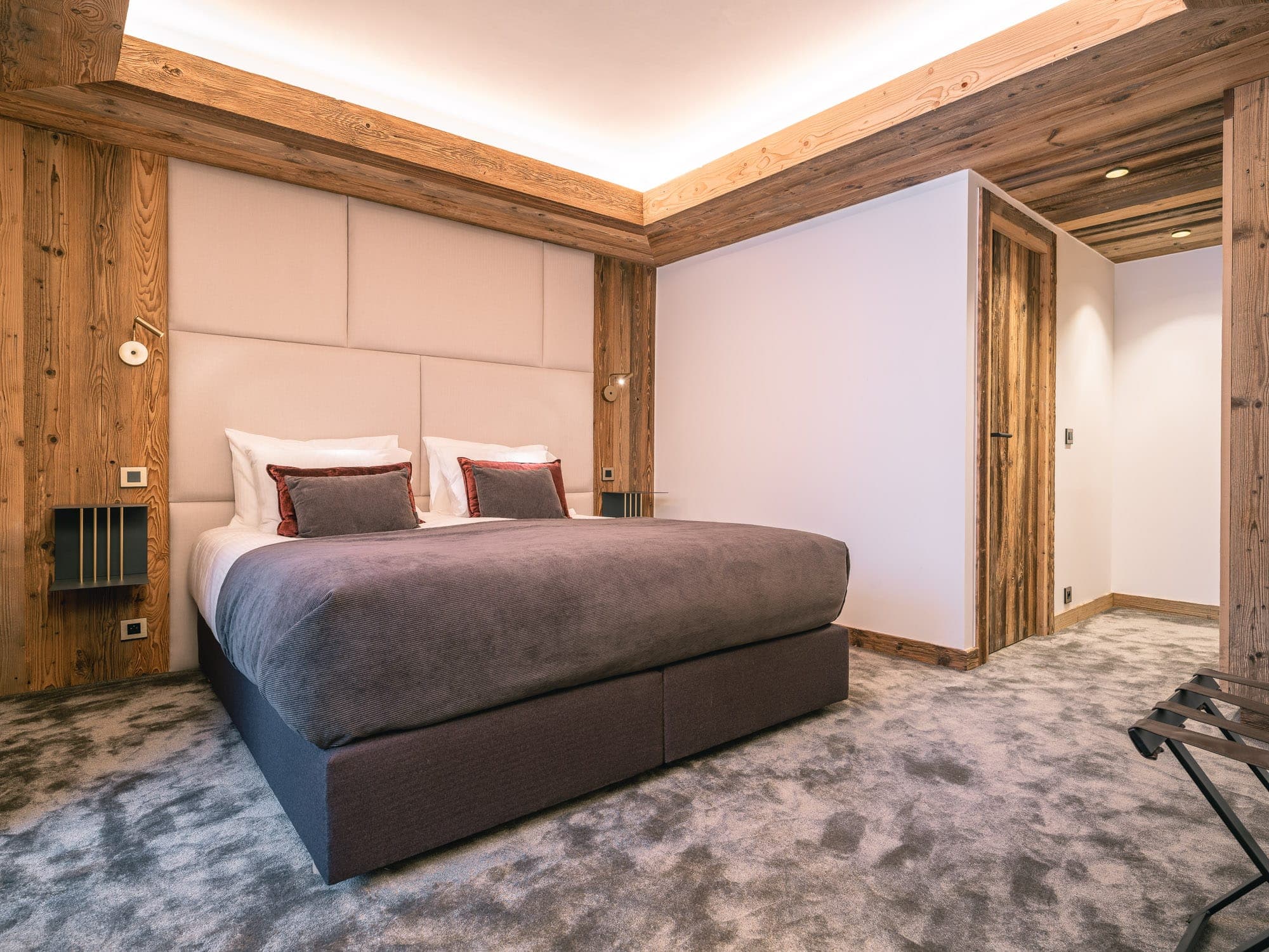 A modern bedroom with warm woodwork includes a king-size bed with brown bedding and white pillows. An upholstered headboard and wall lights add comfort and style. A soft carpet covers the floor, creating a warm ambience.
