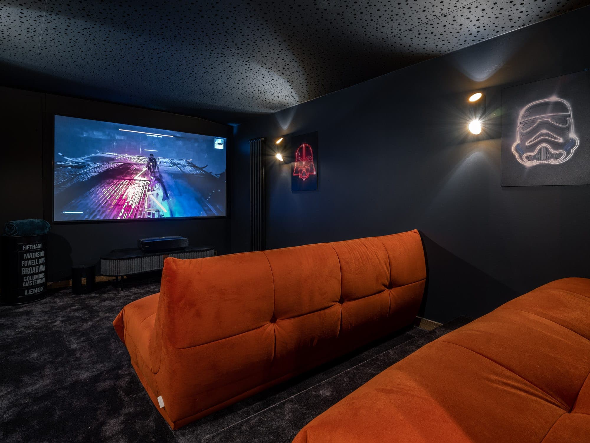 A cosy home cinema with two orange sofas facing a large screen showing a sci-fi scene. The room features subdued lighting, a dark ceiling with star motifs and walls adorned with illuminated artwork, including a Stormtrooper helmet.