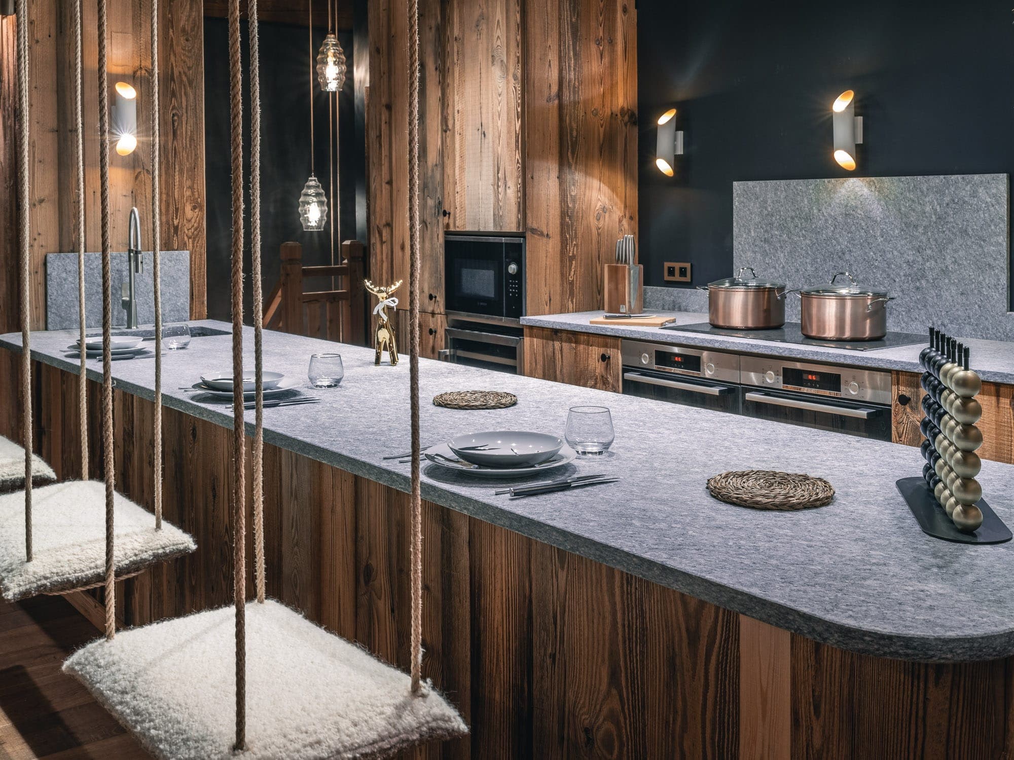 A modern kitchen with rustic wood furniture and flooring. The counter has two swivel seats with cushions. Copper pots and pans are on the stove, accompanied by utensils and glassware. Ambient lighting creates a warm, comfortable atmosphere.