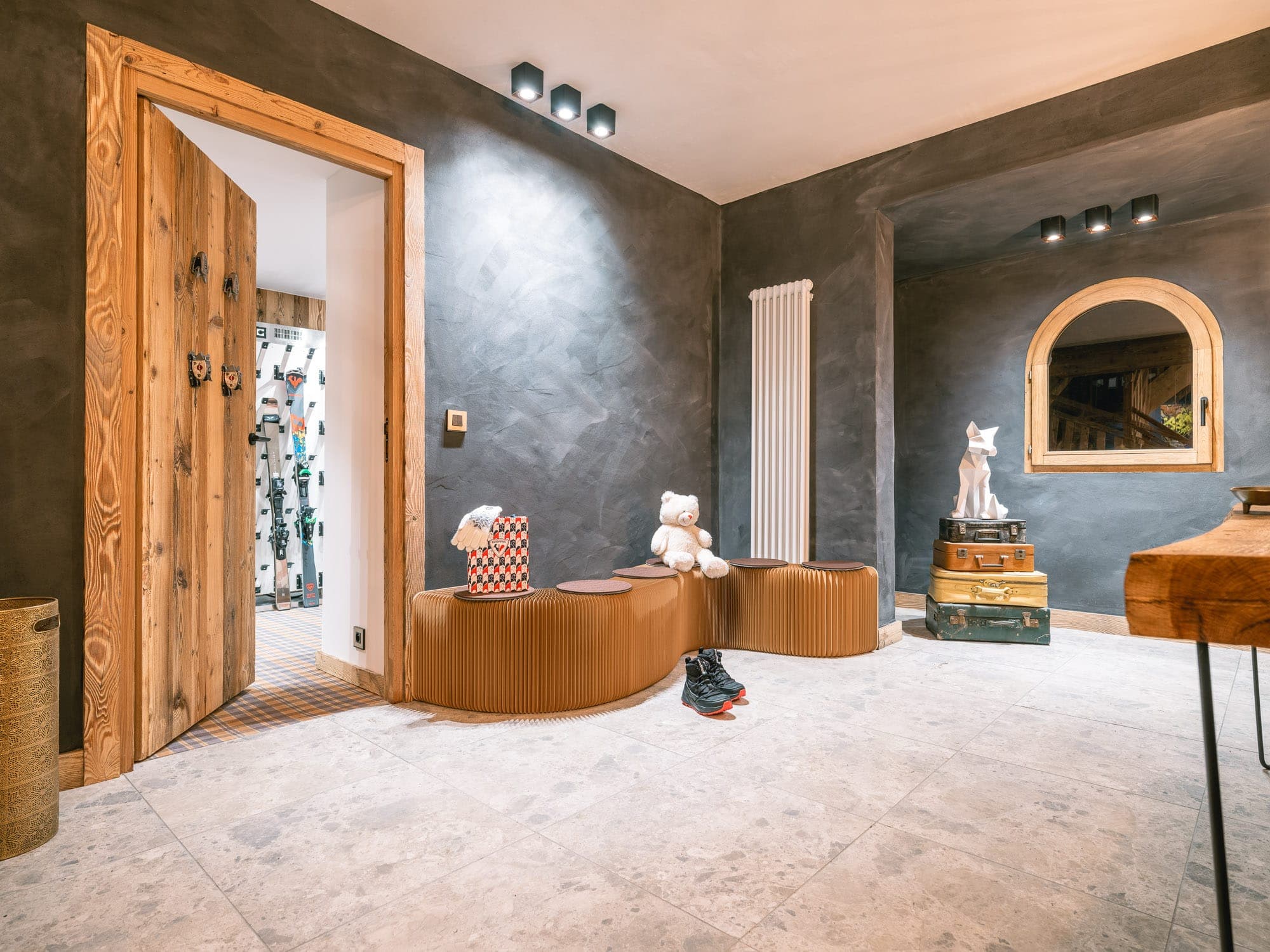 A modern hallway with gray textured walls and light tiles. It features a wooden door, a corrugated wooden bench with plush toys and a shoe on the floor. A mirror with a wooden frame hangs on the wall, above suitcases and a decorative fox.
