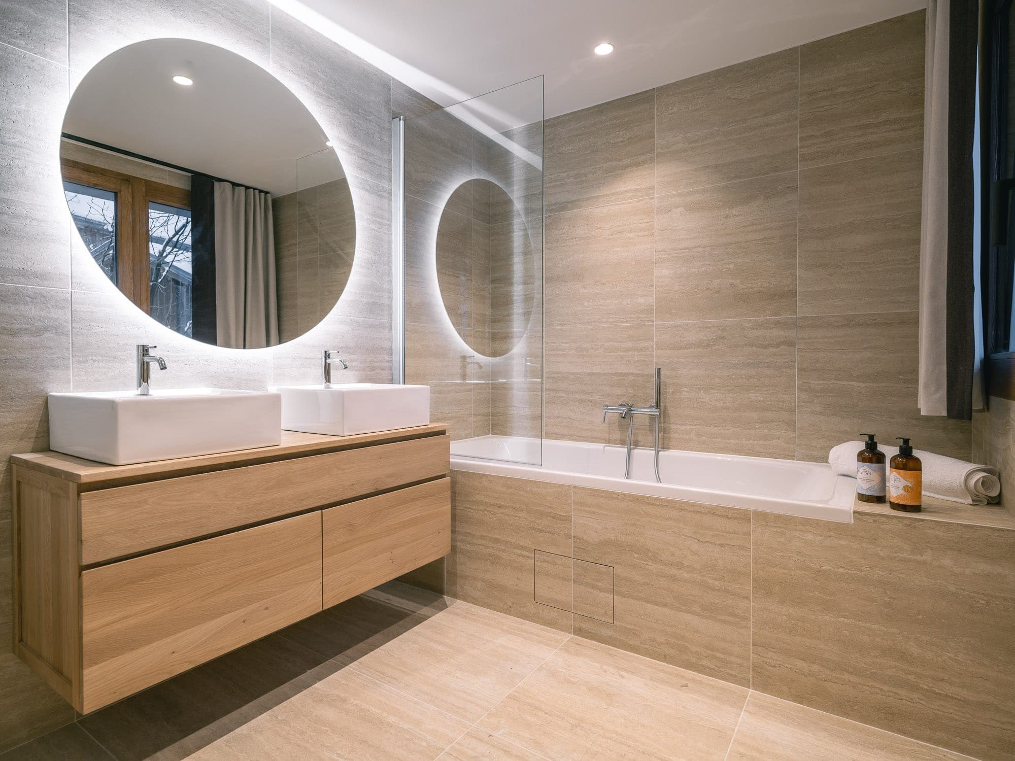 Modern bathroom with light wood cabinet and two rectangular basins. A large round mirror sits above the basins. A bathtub with a glass bath screen is on the right, with bottles and towels on its rim. The room is tiled in beige.