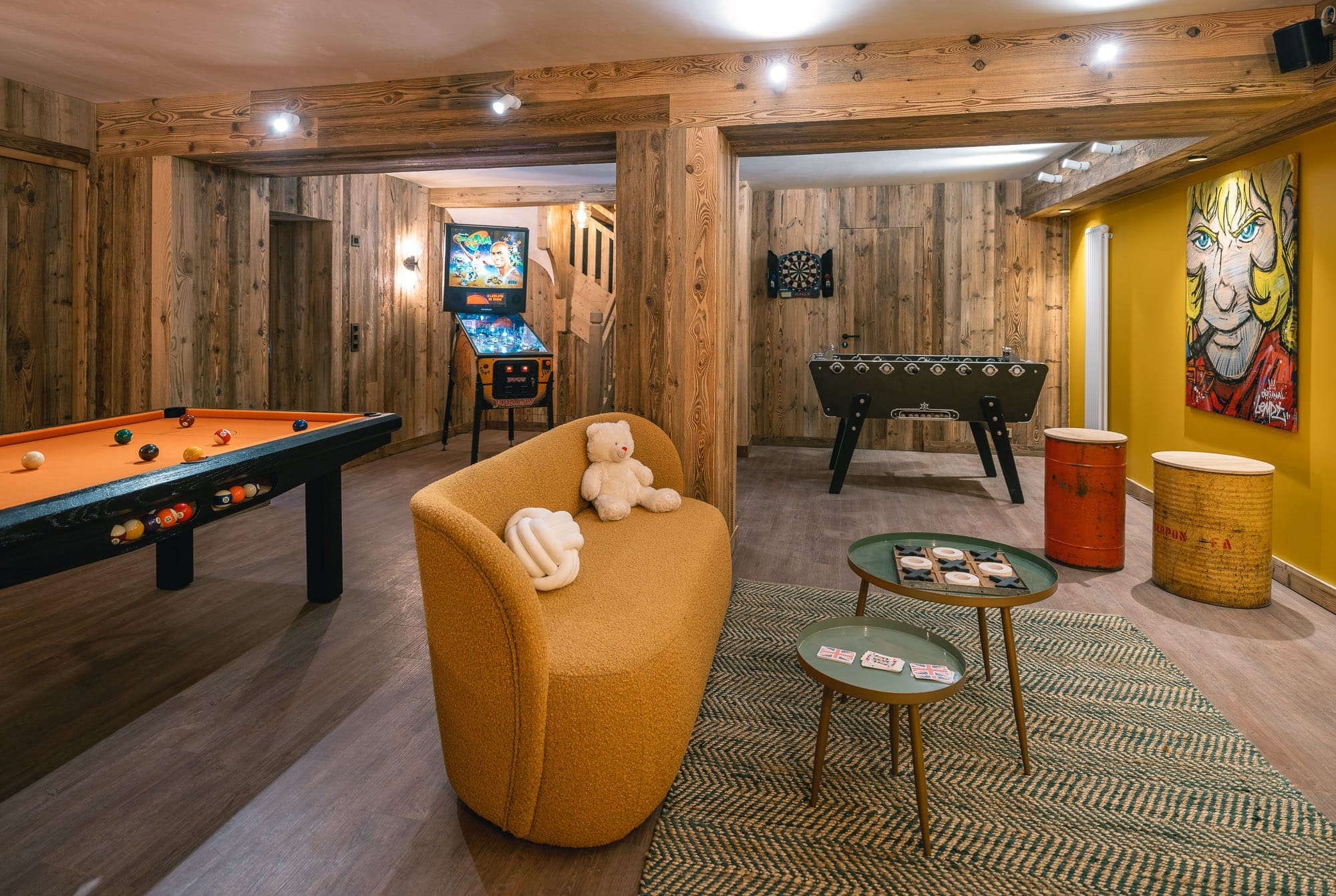 Comfortable games room with paneled walls featuring a pool table, table soccer and pinball machine. A mustard-yellow sofa with plush toys faces two green coffee tables. A dart board and a colorful painting adorn the walls. A woven rug completes the decor.