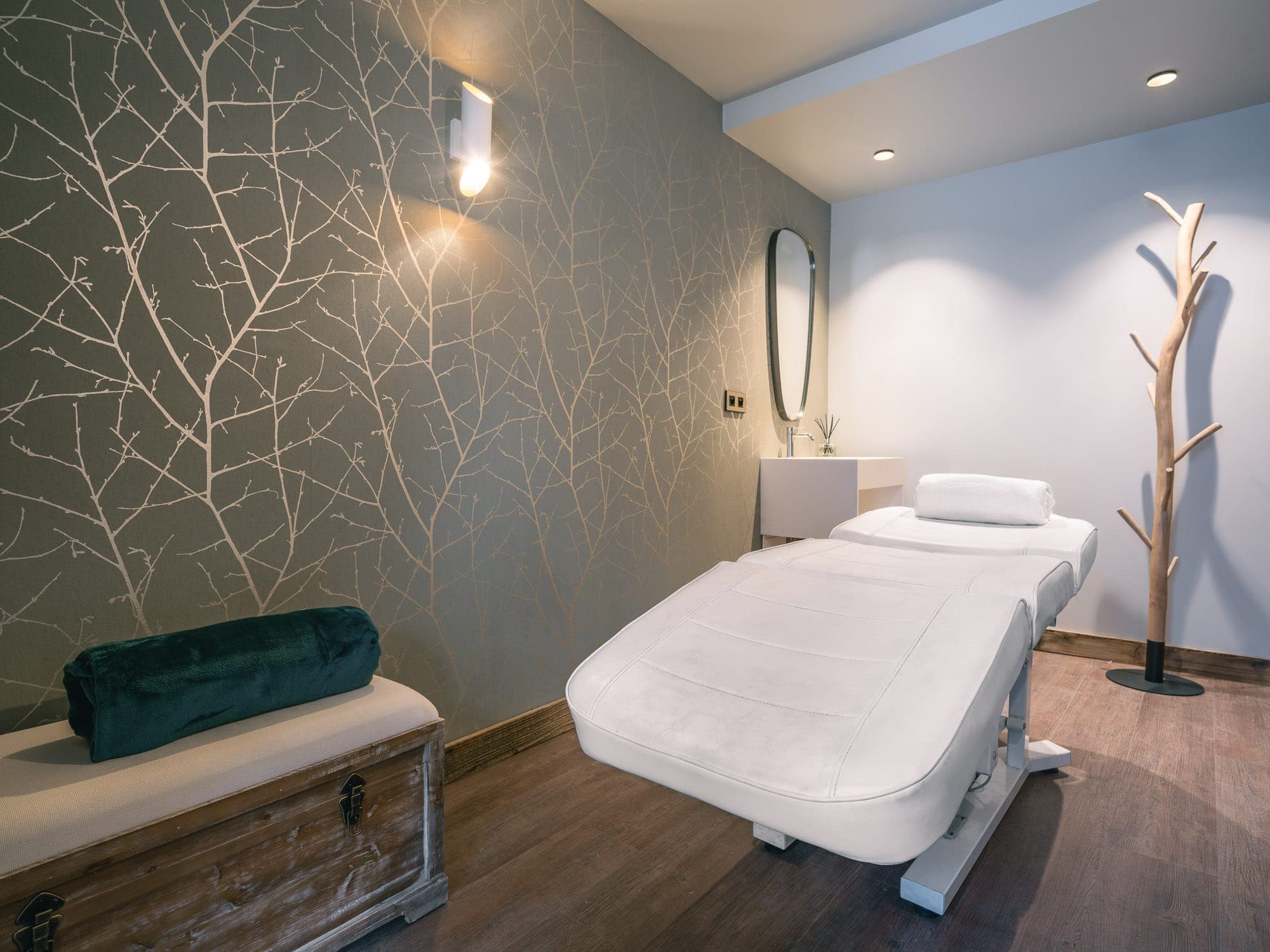 A serene spa room with a white massage table with a pillow, soft lighting and a bench with a green cushion. The walls have a subtle branch design and a unique tree-shaped coat rack stands in the corner.