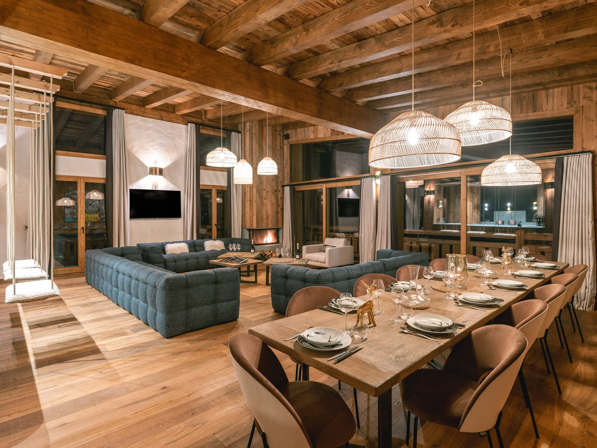 Spacious rustic living and dining room with wooden beams and floors. Features a long dining table, soft blue sofas, cozy fireplace and large windows. Hanging lamps create a warm ambience.