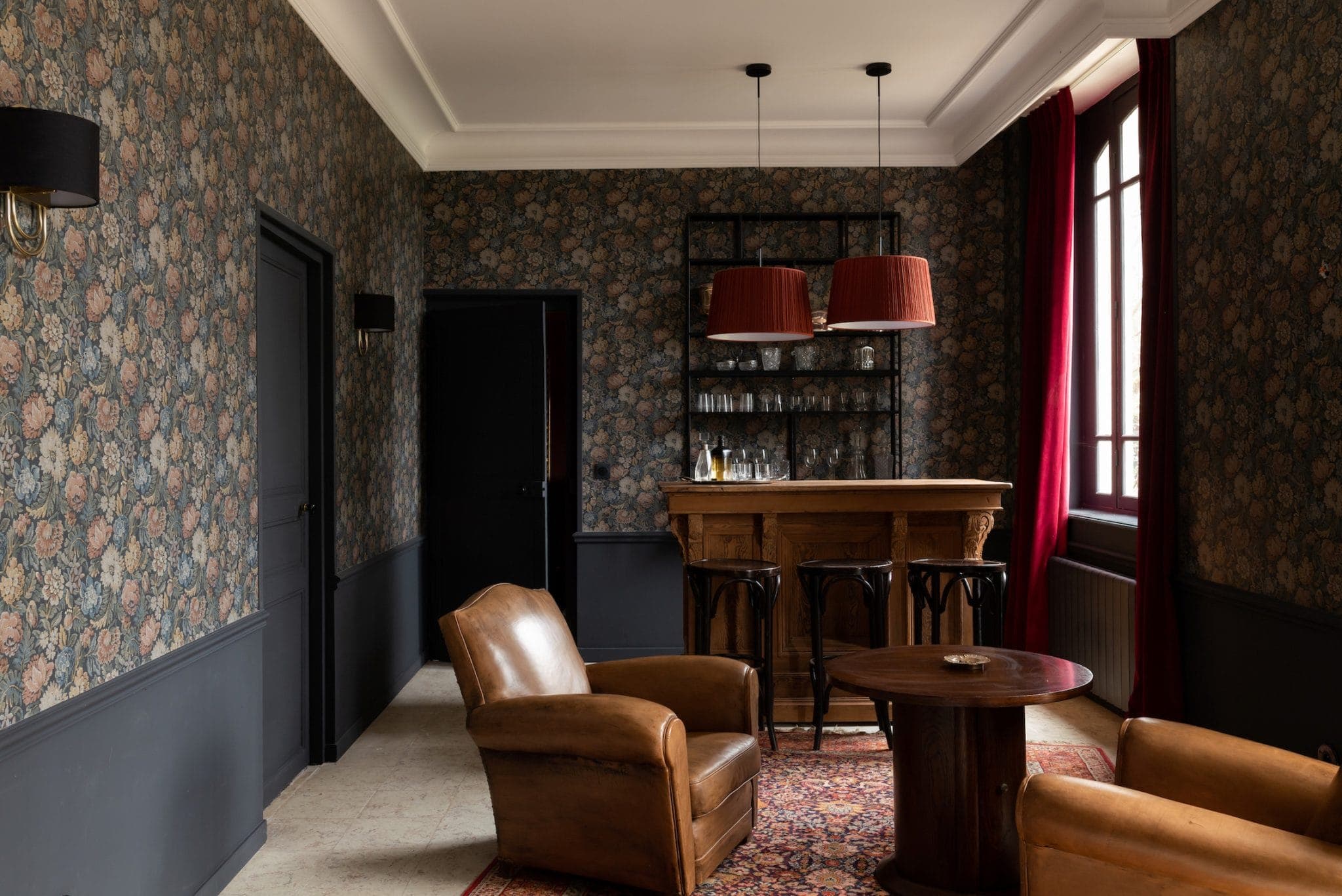 Une salle confortable est dotée de fauteuils en cuir, d'une table en bois et d'un tapis à motifs. Un petit bar avec tabourets et verres se trouve dans le coin. Les murs sont ornés de papier peint fleuri et de grandes fenêtres avec des rideaux rouges laissent entrer la lumière naturelle.