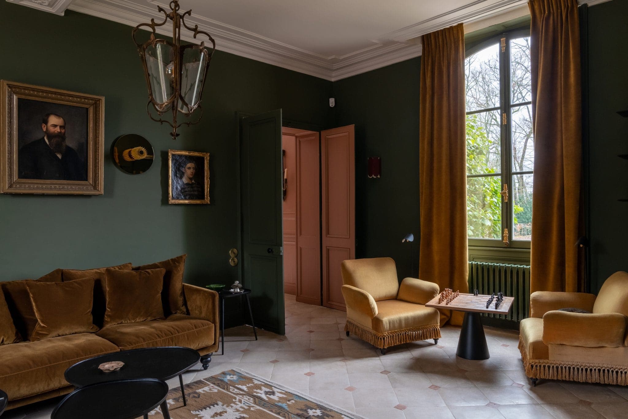 Un salon cosy aux murs vert foncé, une grande fenêtre avec des rideaux moutarde et une décoration vintage. Il y a des fauteuils couleur moutarde, un canapé et un jeu d'échecs sur la table, avec des portraits encadrés sur les murs.