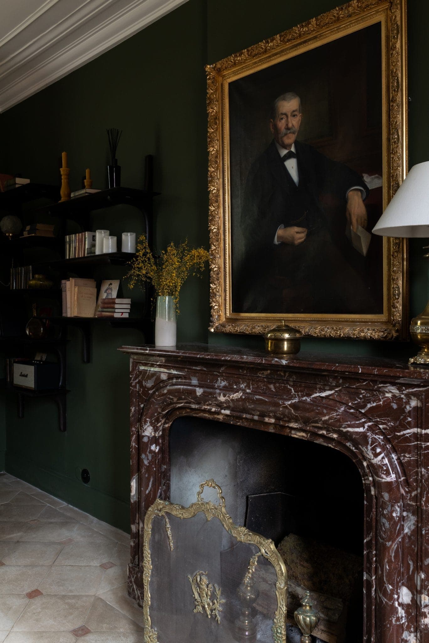 Chambre élégante aux murs vert foncé et au grand portrait d'un homme en costume noir. Une cheminée en marbre sous le tableau, ornée d'un paravent à cadre doré. Des étagères avec des livres et des décorations se trouvent à gauche, et une lampe blanche sur le manteau.
