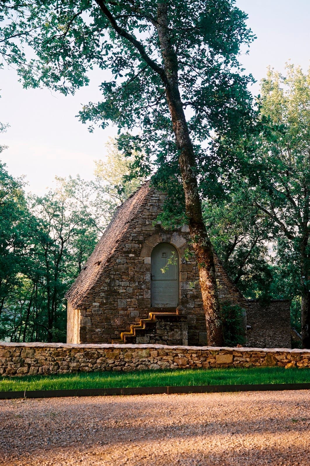 Une petite maison en pierre au toit triangulaire, conçue par Sapiens Architectes, est nichée au milieu de grands arbres. On y accède par un petit escalier en pierre. La lumière du soleil filtre à travers la verdure, créant des ombres tachetées sur le sol. Un mur de pierre court devant.