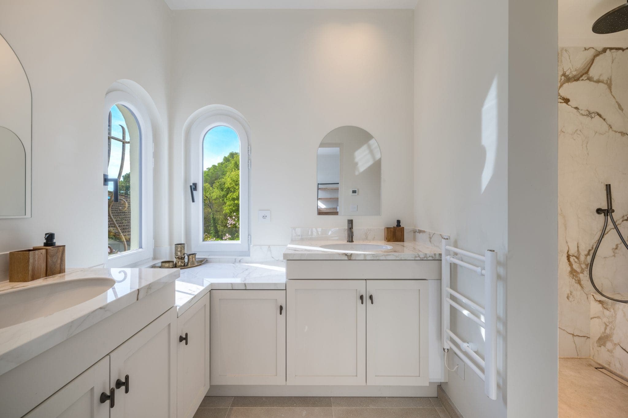 Modern bathroom with white cabinetry, marble countertops and arched windows. A deep bathtub is nestled beneath the windows, and a walk-in shower with marble tiling is on the right. Natural light fills the room, enhancing the minimalist design.