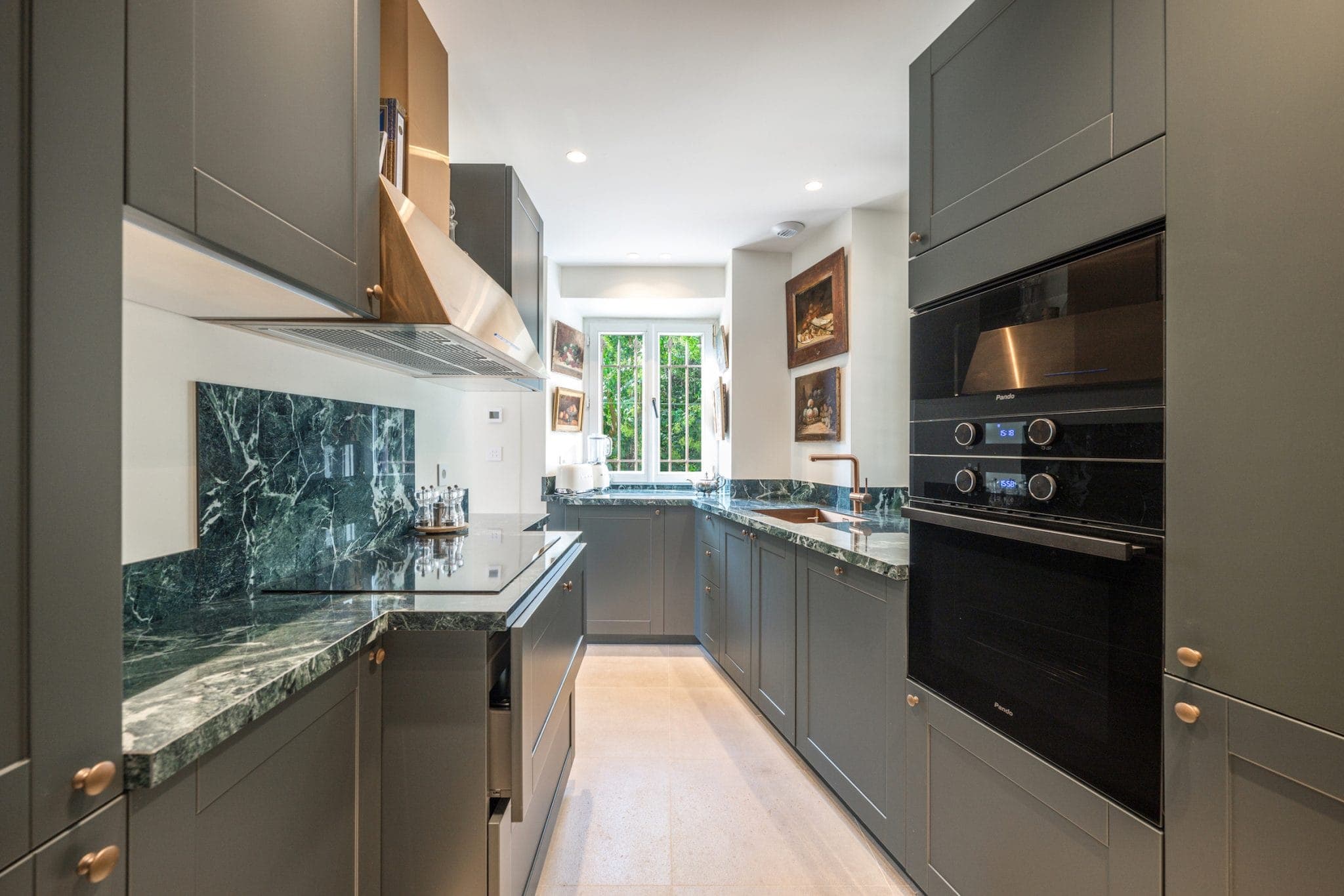 A modern kitchen with green cabinetry, marble countertops and a black double oven. A window at the back lets in natural light, illuminating the artwork on the walls. The floor is in light-colored tiles.
