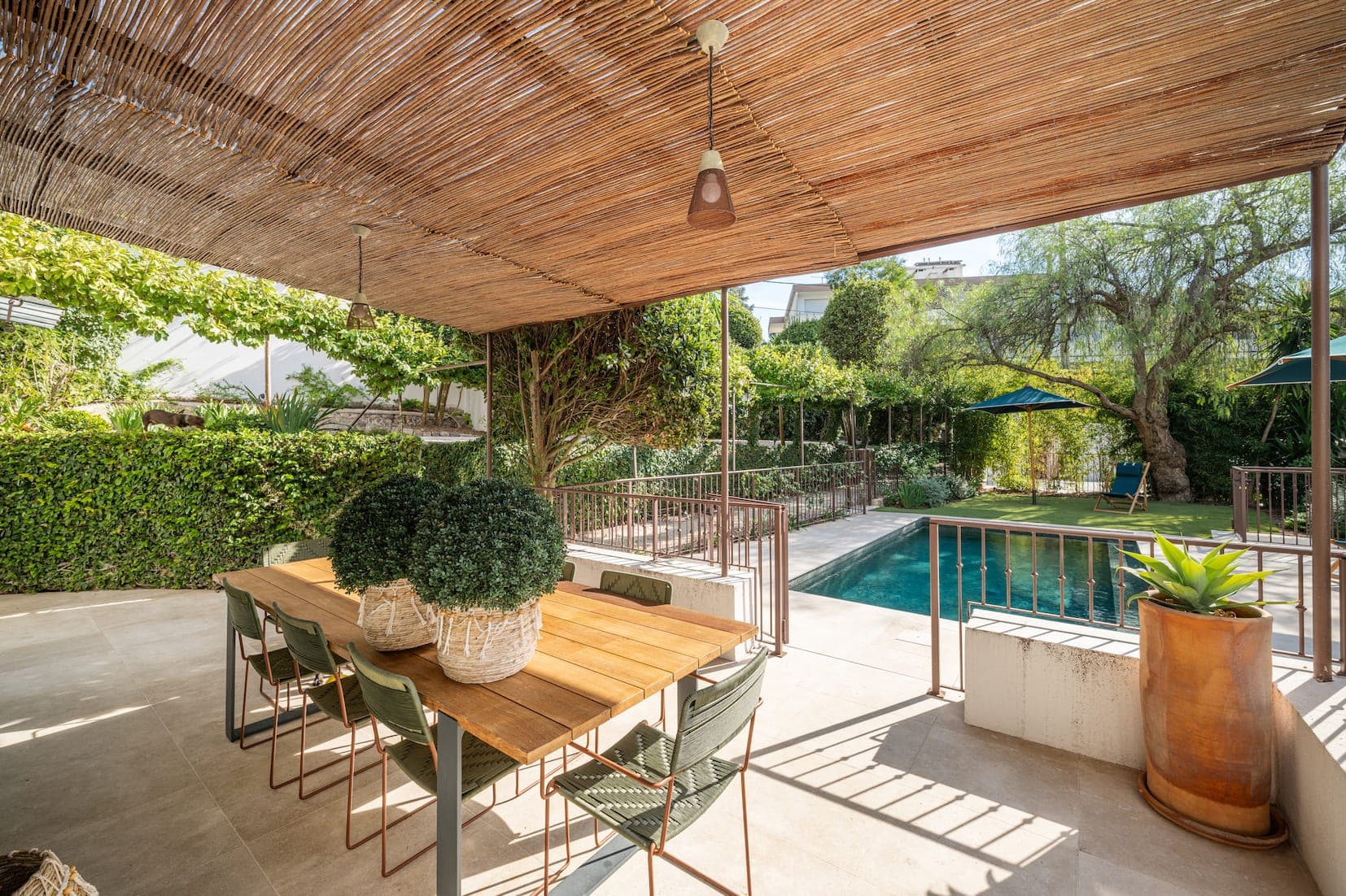 Outdoor terrace with wooden table and green chairs under a bamboo canopy. The space overlooks a swimming pool surrounded by lush vegetation and trees. Potted plants adorn the space, creating a tranquil atmosphere steeped in nature.