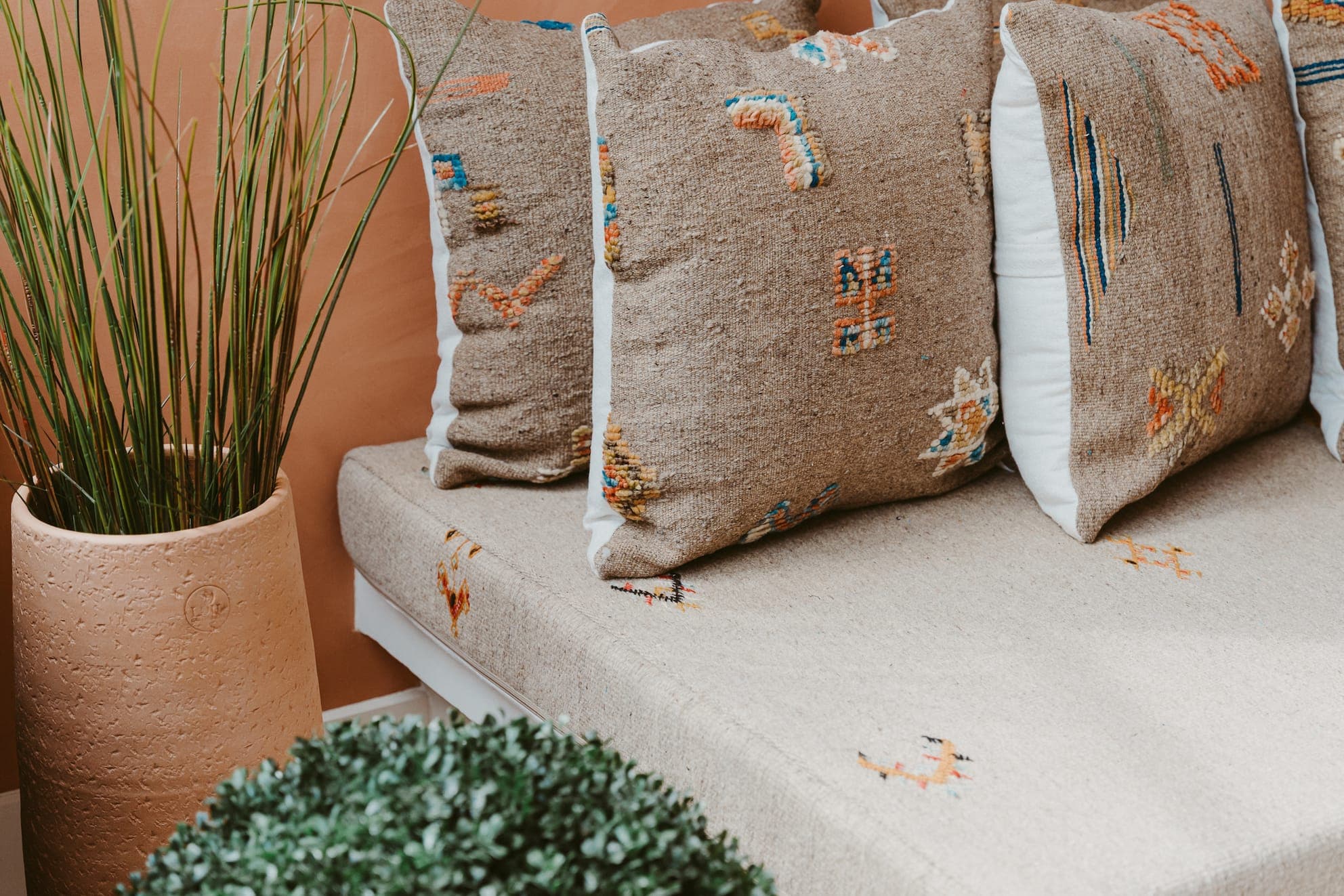 A comfortable sitting area with an upholstered beige bench adorned with decorative cushions in earthy tones. A large terracotta pot holds tall green grass and a small round bush decorates the foreground. The décor exudes a warm, natural ambience.