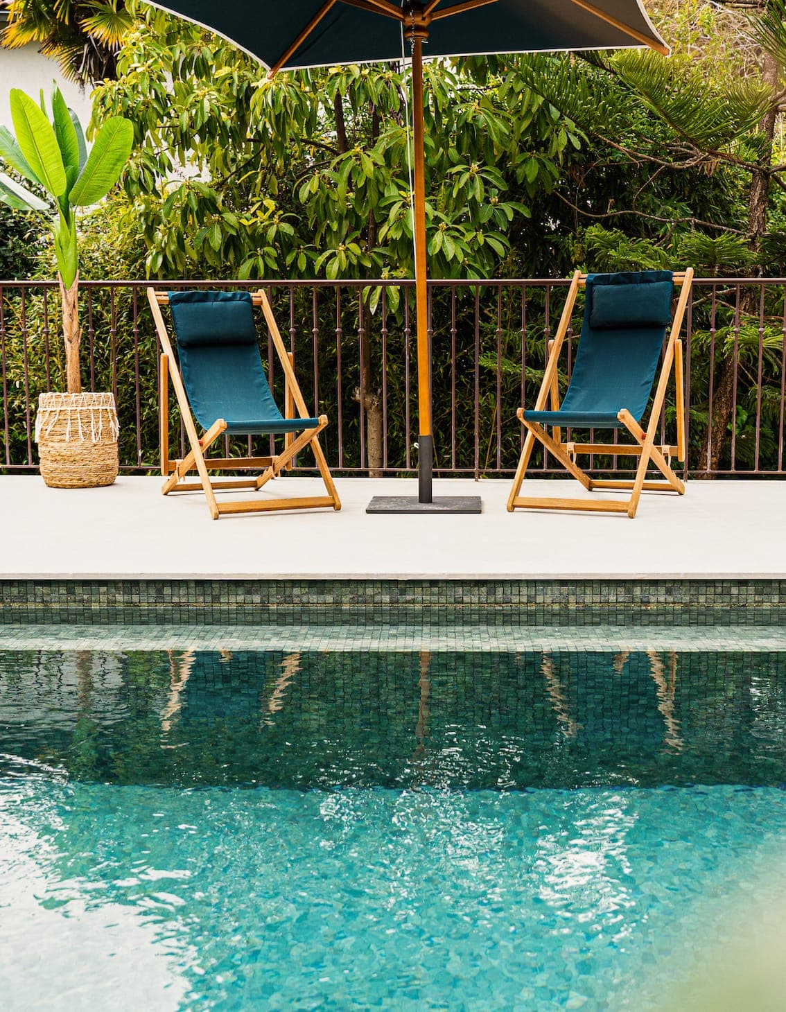 Two wooden loungers with dark cushions sit beneath a large parasol on a poolside terrace. A woven basket and lush green plants are in the background, creating a serene, tropical atmosphere. The pool reflects the greenery.