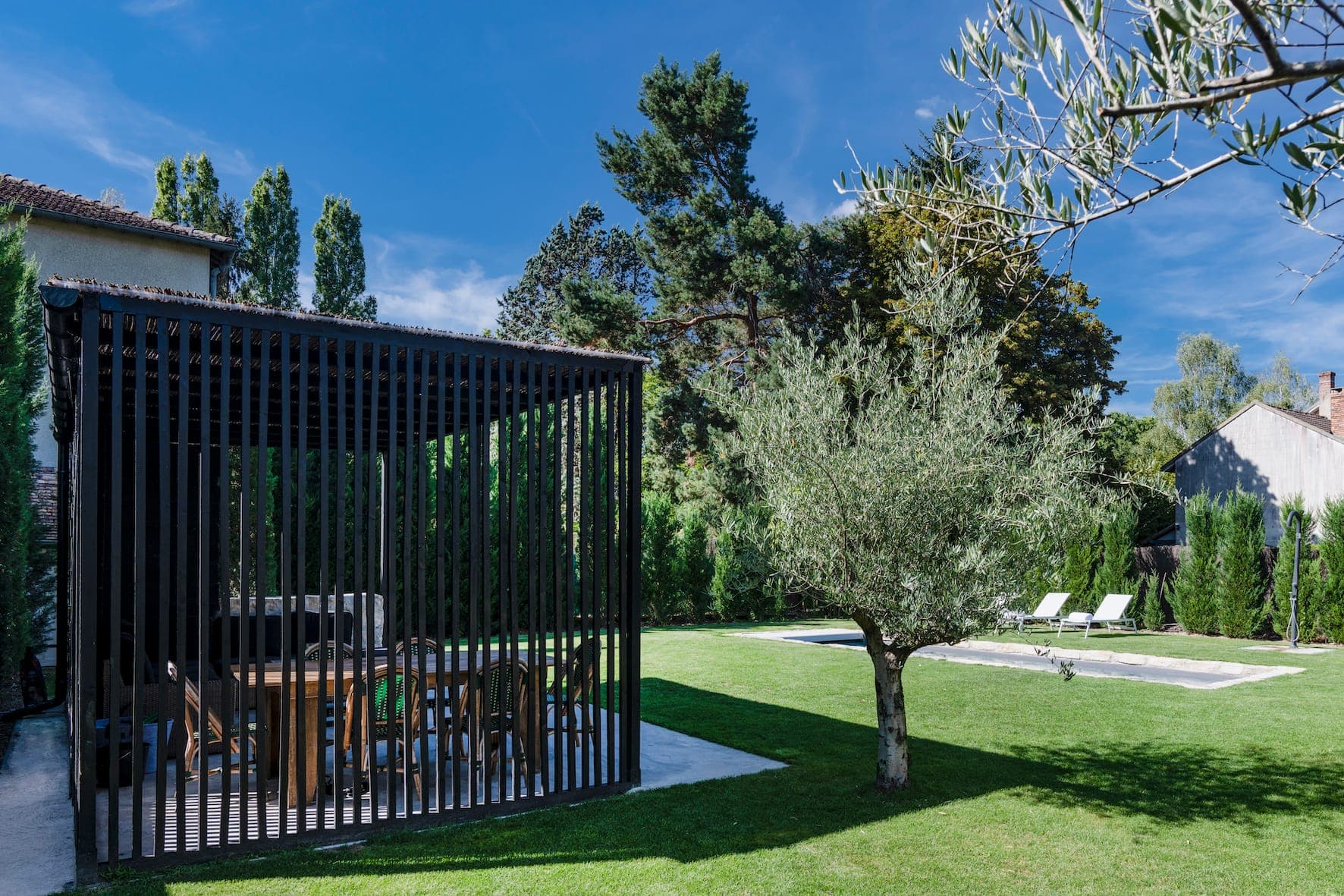 A terrace with a wooden slatted fence includes a dining table and chairs. It is set in a lush green lawn with a tree nearby and a swimming pool in the background. The sky is clear and blue, and the area is surrounded by tall trees and shrubs.