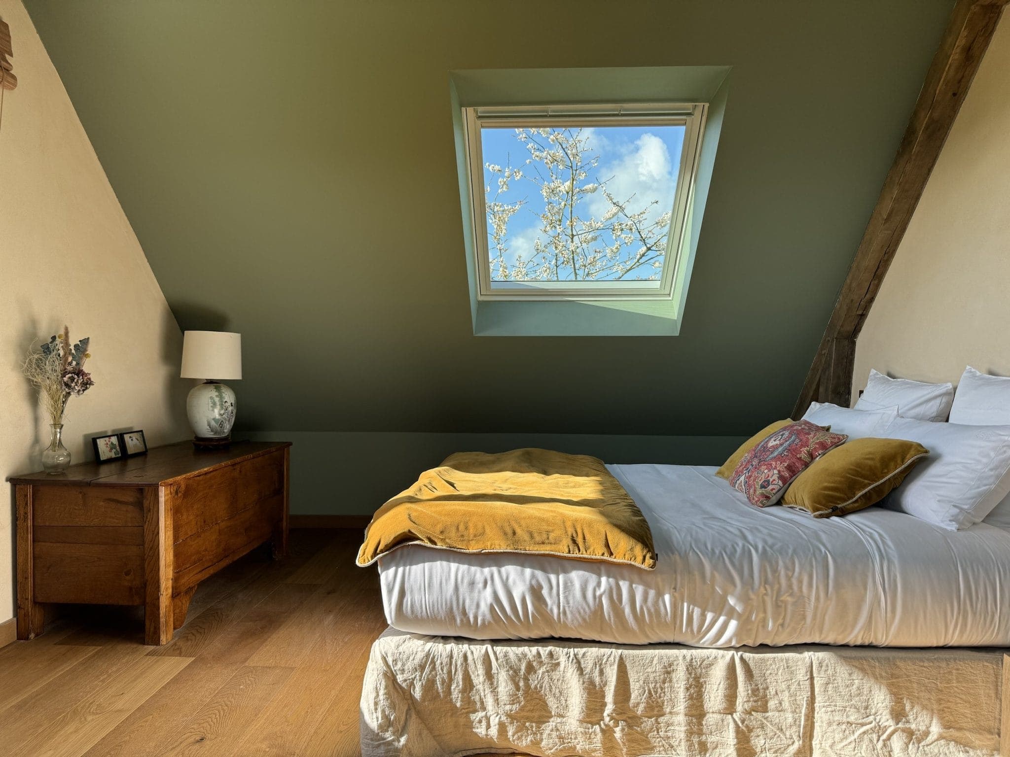 Comfortable attic room with a bed with white and mustard bedding, adjacent to a wooden chest. A skylight above reveals a blue sky. A small lamp and framed photos sit on the chest, while a rustic beam accentuates the sloping green ceiling.