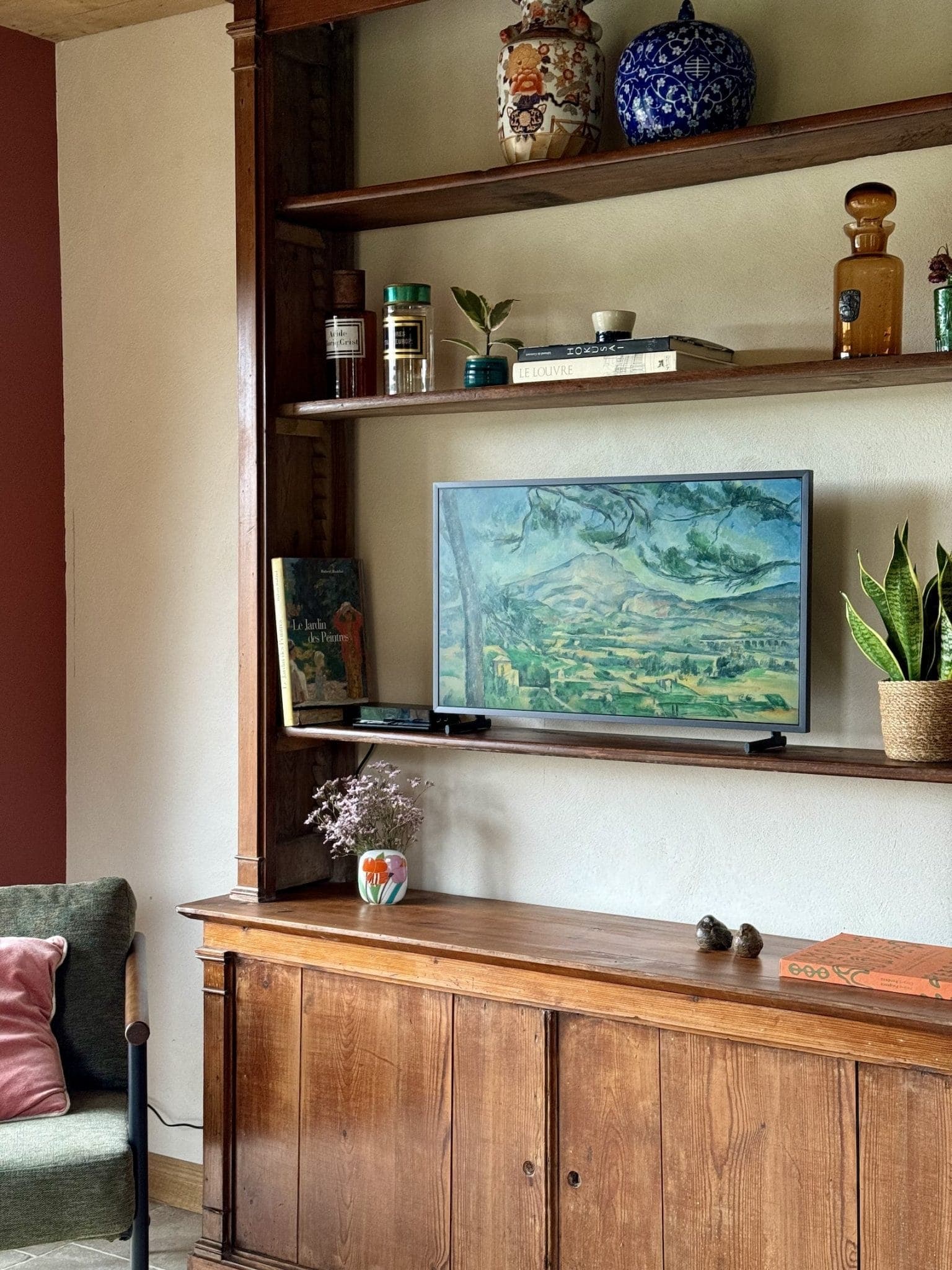 A comfortable living room features a wooden bookcase with decorative objects, books and a small TV displaying a picturesque painting. A potted plant and vase adorn the cabinet below, with a green chair and red cushion in the corner.