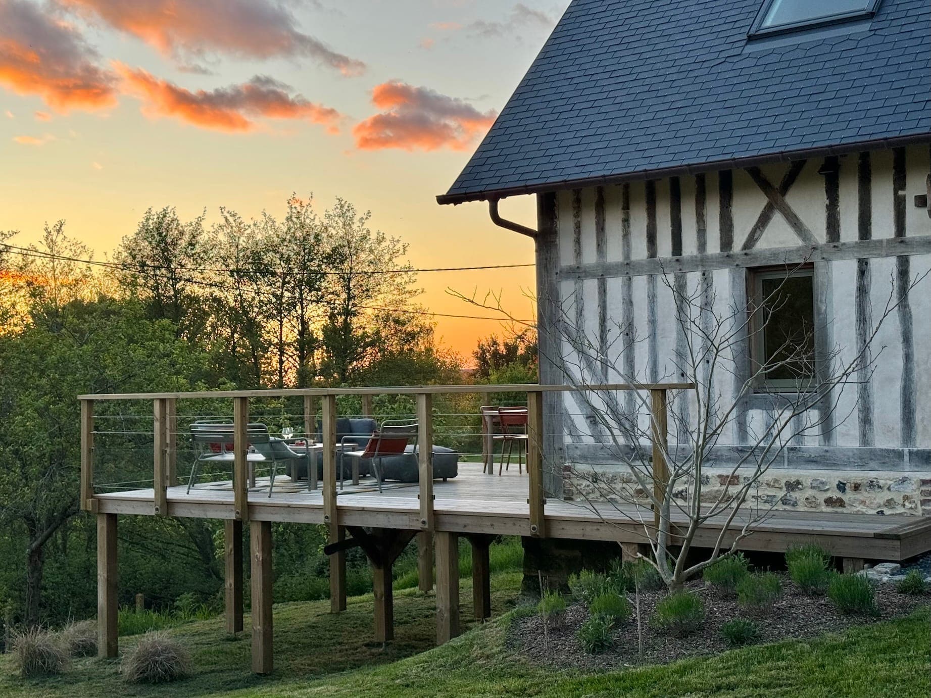 Une maison à ossature bois avec une terrasse en bois surélevée surplombe une cour herbeuse au coucher du soleil. La terrasse est dotée de balustrades en verre et de meubles d'extérieur. Le ciel est peint de teintes orange et roses, et les arbres entourent la zone.