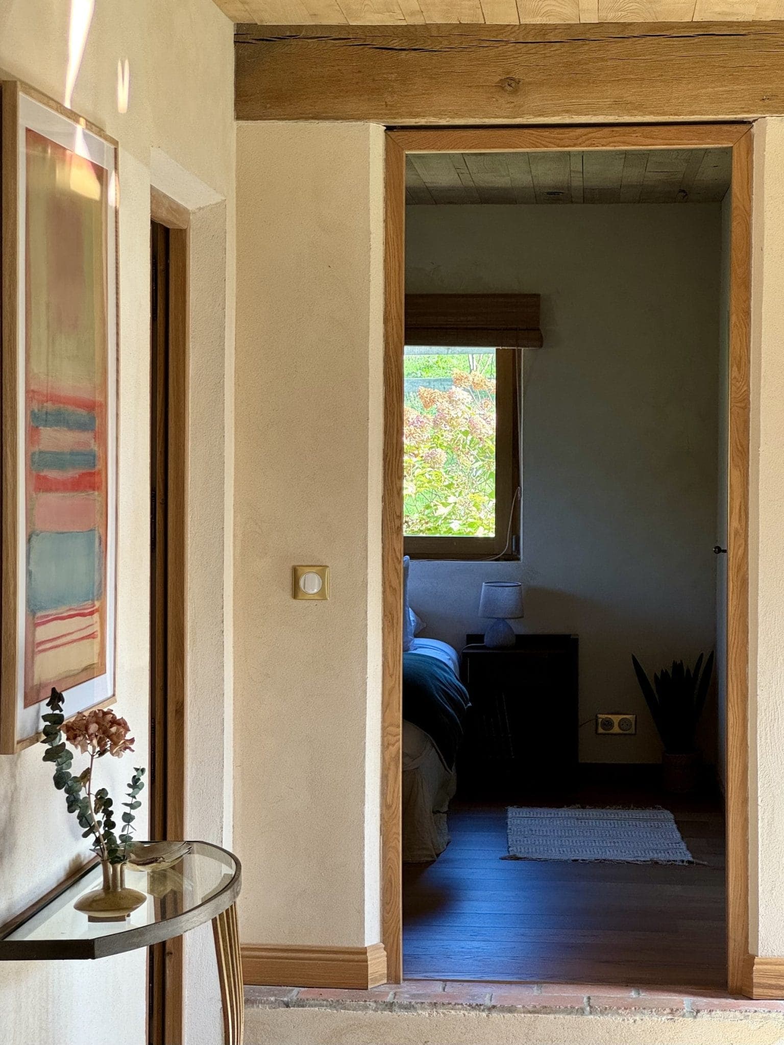 View through a hallway with a wooden frame leading to a softly lit bedroom. On the left wall is a colorful abstract painting. A small glass table below holds a vase with flowers. A window in the bedroom offers a glimpse of the greenery outside.
