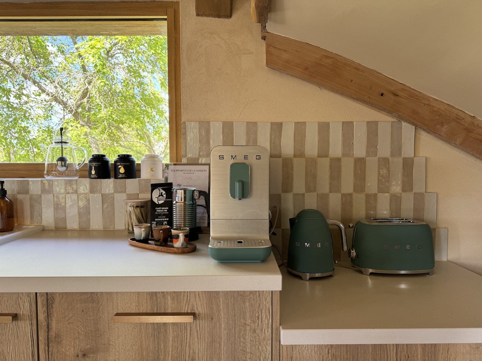 A kitchen counter with a Smeg coffee machine, kettle and toaster in green. A wooden tray holds coffee essentials. A ceramic-tiled backsplash, wooden cabinets, a window and sunlight on the leaves create a warm atmosphere.
