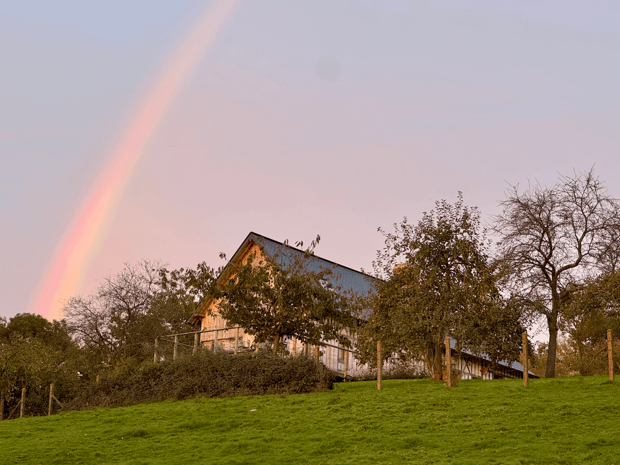 A rustic blue-roofed barn is nestled among trees on a grassy hillside. A vibrant rainbow crosses the sky in the early evening, adding a colorful backdrop to the peaceful rural scene.