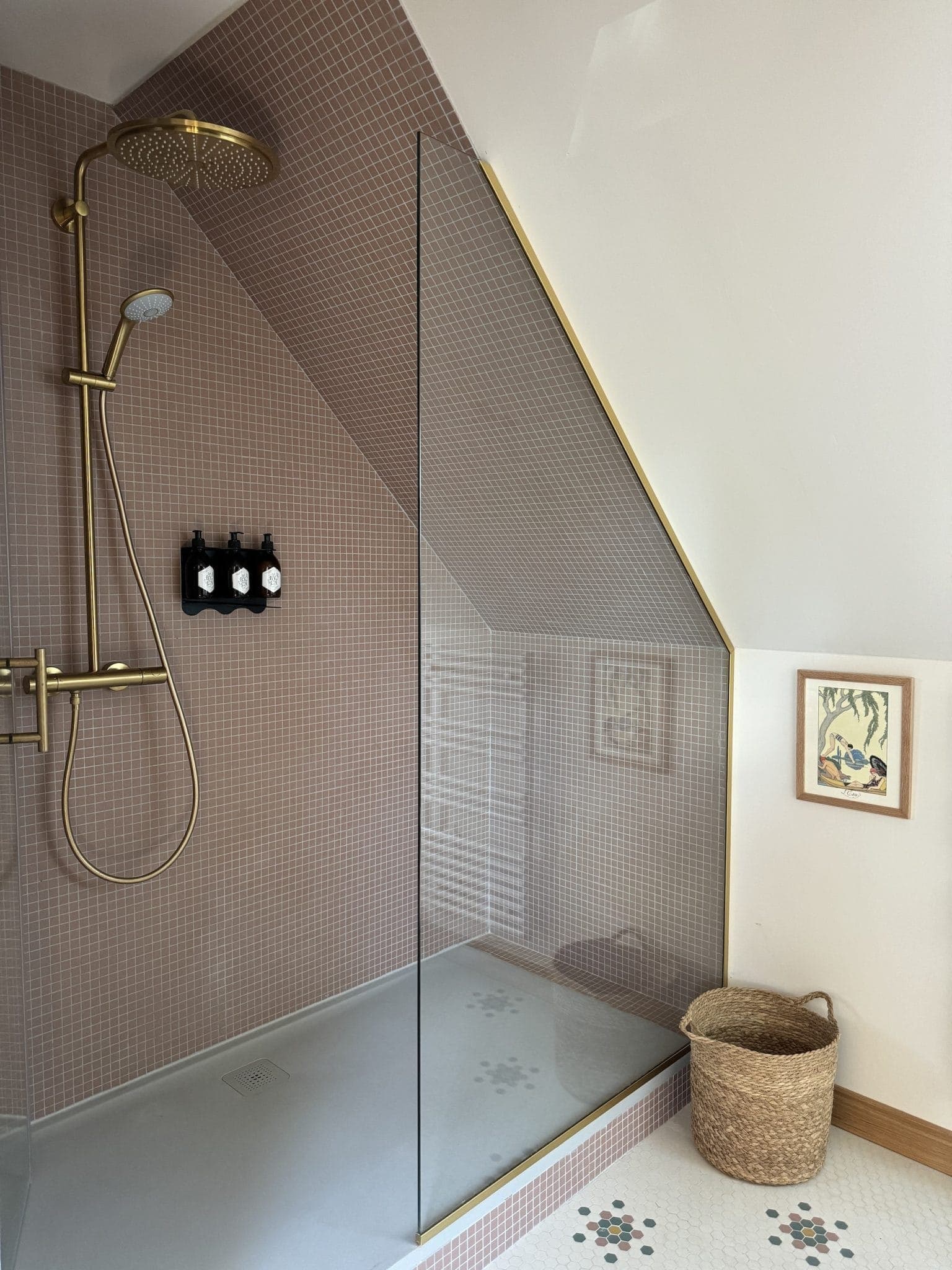 A modern shower with rainfall showerhead and gold accessories is set against a pink tiled wall. A glass panel separates the shower from the rest of the bathroom. A wicker basket and artwork adorn the floor and wall outside the shower.