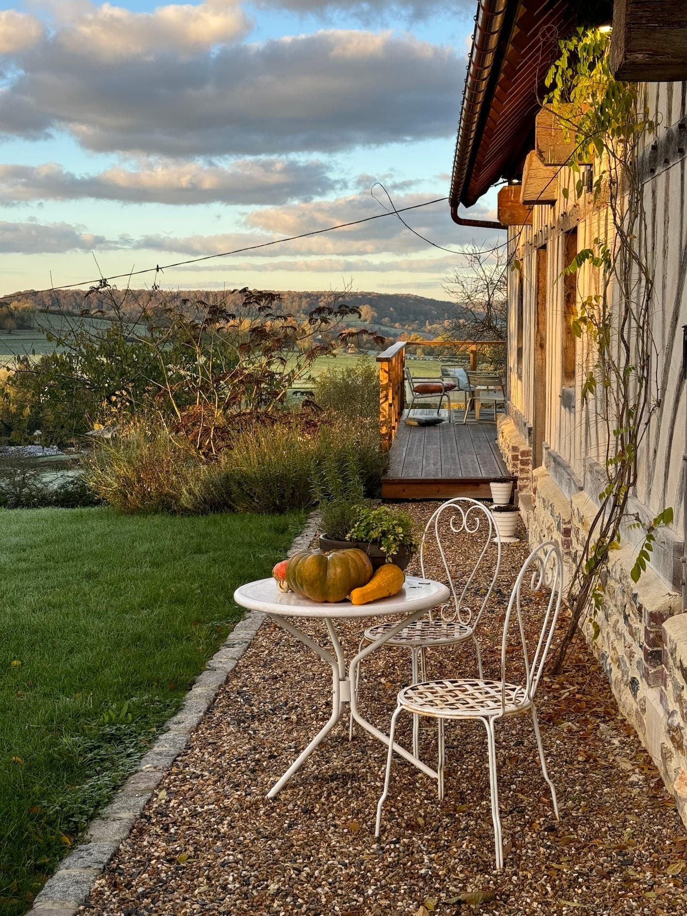 A rustic outdoor setting with a white metal table and chairs on a gravel terrace, adorned with pumpkins. The scene overlooks a distant view of hills and greenery under a partly cloudy sky. Ivy grows on the nearby wooden house.