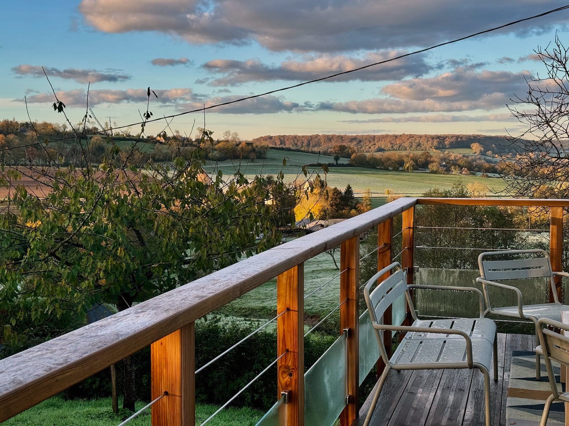 Panoramic views from a wooden terrace with metal balustrades overlooking a lush, rolling landscape. Chairs are arranged on the terrace. The sky is partly cloudy and the low sun casts a warm light over the fields and trees, highlighting the colors of autumn.
