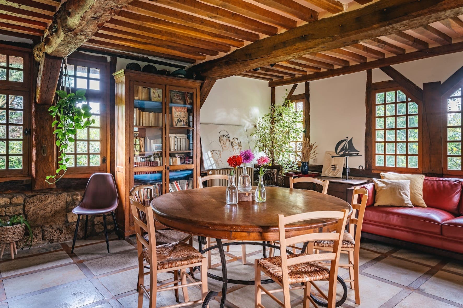 Un coin repas chaleureux avec une table ronde en bois et des chaises. Il y a un canapé rouge, une bibliothèque et des plantes en pot. Des poutres en bois apparentes et un mur en pierre ajoutent un charme rustique. De nombreuses fenêtres laissent entrer la lumière naturelle, créant une atmosphère chaleureuse.