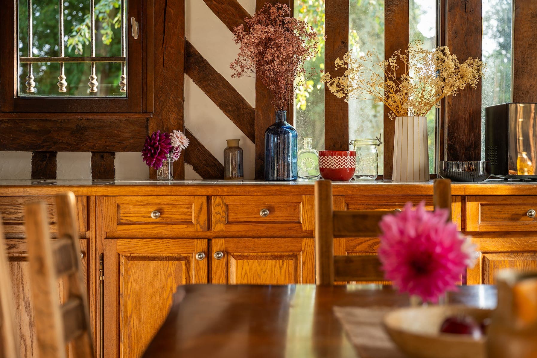 Une cuisine chaleureuse avec des armoires en bois et une décoration rustique. Diverses fleurs séchées dans des vases sont posées sur le comptoir près de la fenêtre. Une table en bois avec des chaises est au premier plan, avec un vase avec des fleurs roses. La lumière du soleil filtre à travers la fenêtre.