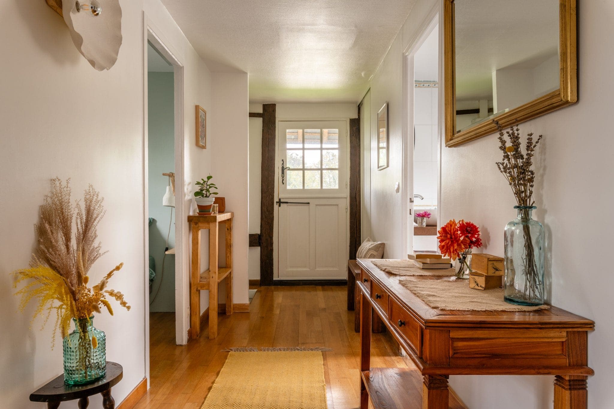 Un couloir chaleureux avec parquet en bois, avec une console ornée de livres, un vase bleu et des fleurs rouges. Un miroir mural est suspendu au-dessus de la table. Une petite table avec une plante se trouve près de la porte blanche en arrière-plan. La lumière naturelle illumine l'espace.