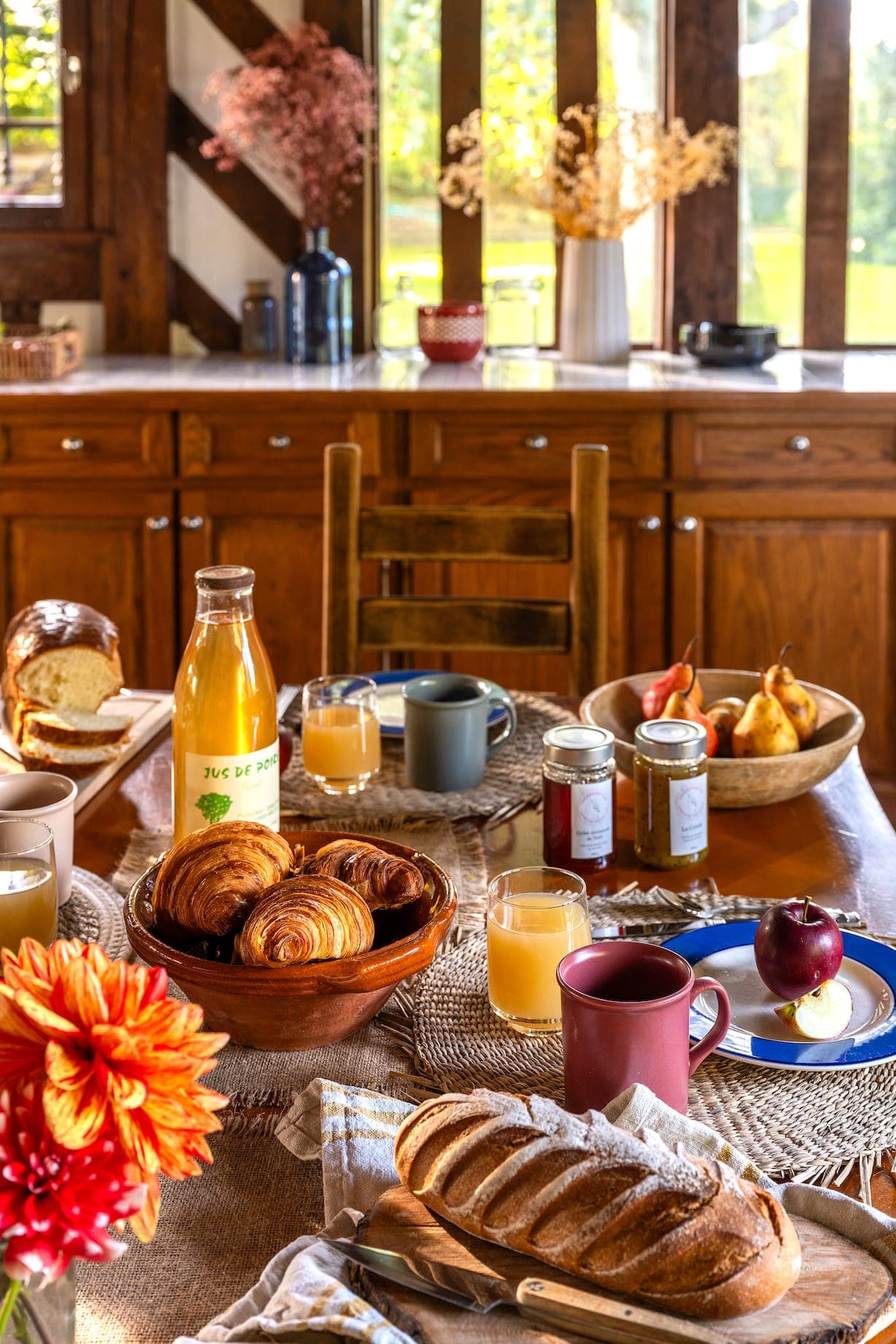 Une cuisine confortable est dotée d'une table en bois avec une variété d'articles pour le petit-déjeuner : un pichet de jus d'orange, des croissants, de la confiture, des poires, du pain et des tasses. La lumière du soleil pénètre par la fenêtre, mettant en valeur les fleurs colorées et la décoration rustique.