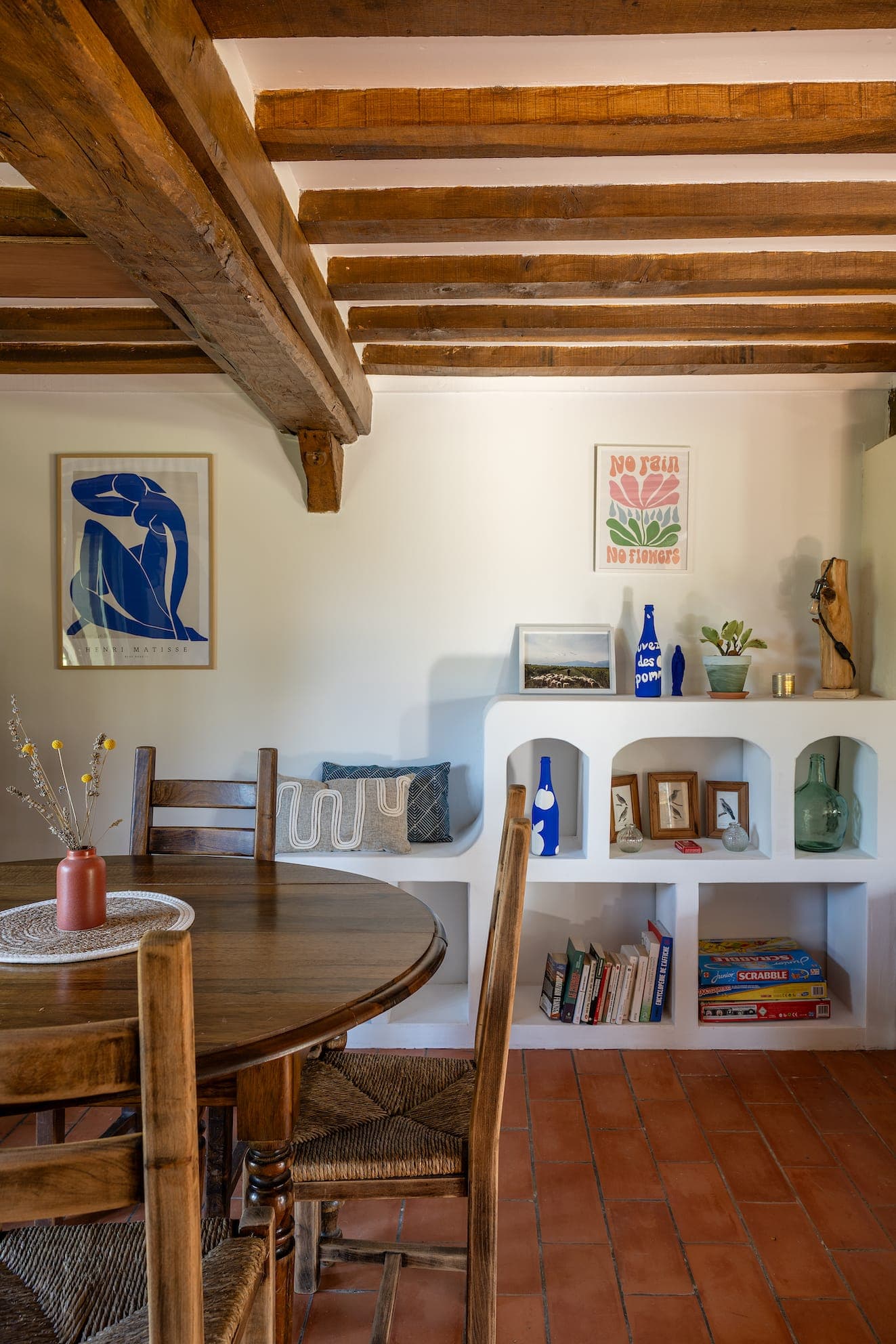 Une pièce chaleureuse avec poutres apparentes et sol en carrelage en terre cuite. Elle comprend une table à manger ronde en bois avec des chaises. Des étagères blanches présentent des livres, des œuvres d'art et des plantes. Deux affiches colorées ornent les murs blancs, ajoutant de la chaleur à l'espace.