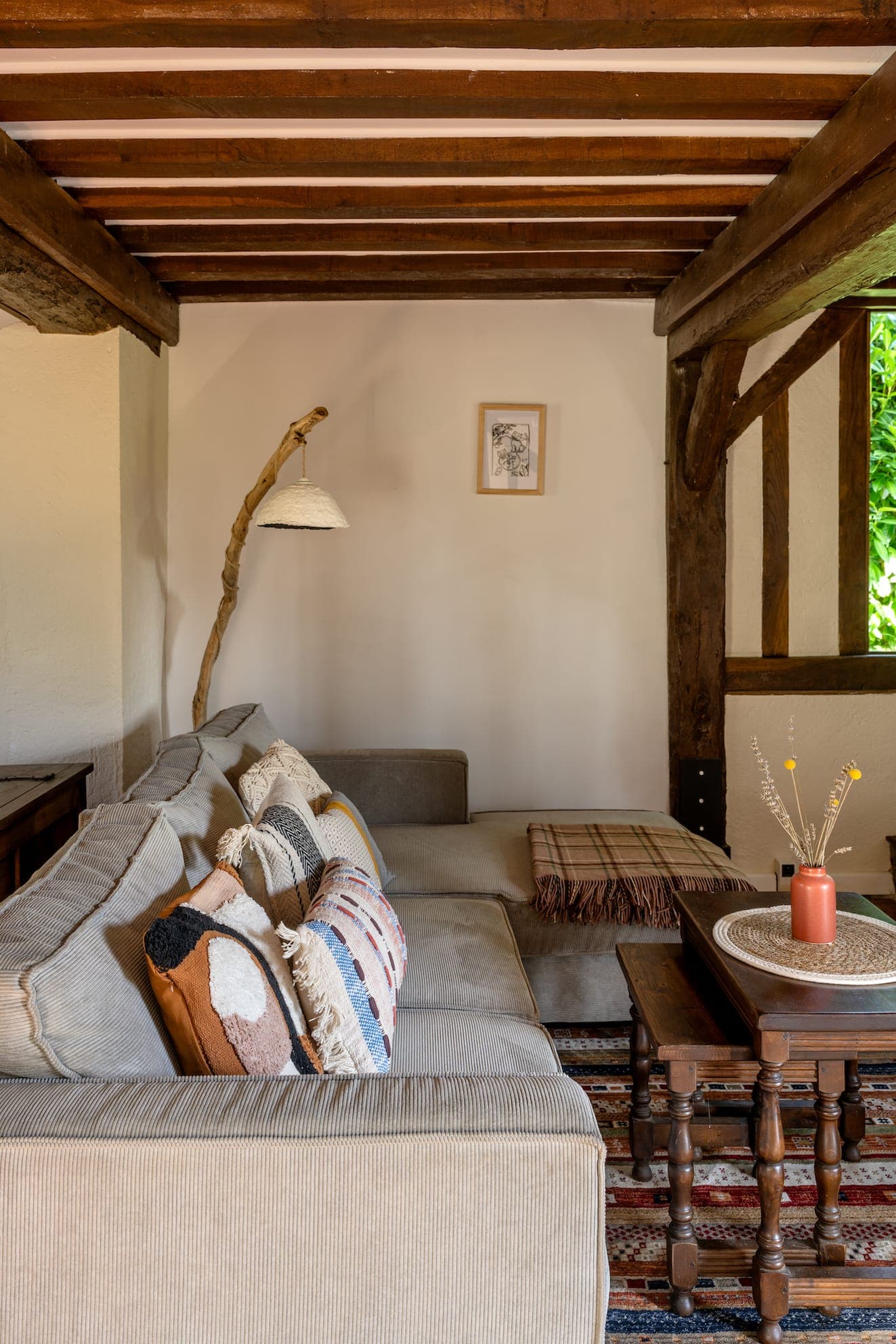 Salon confortable avec un canapé d'angle rayé orné de coussins à motifs. Une table basse en bois rustique accueille un vase avec des fleurs séchées. La pièce est dotée de poutres en bois, d'un lampadaire unique et d'un petit tableau encadré sur le mur.