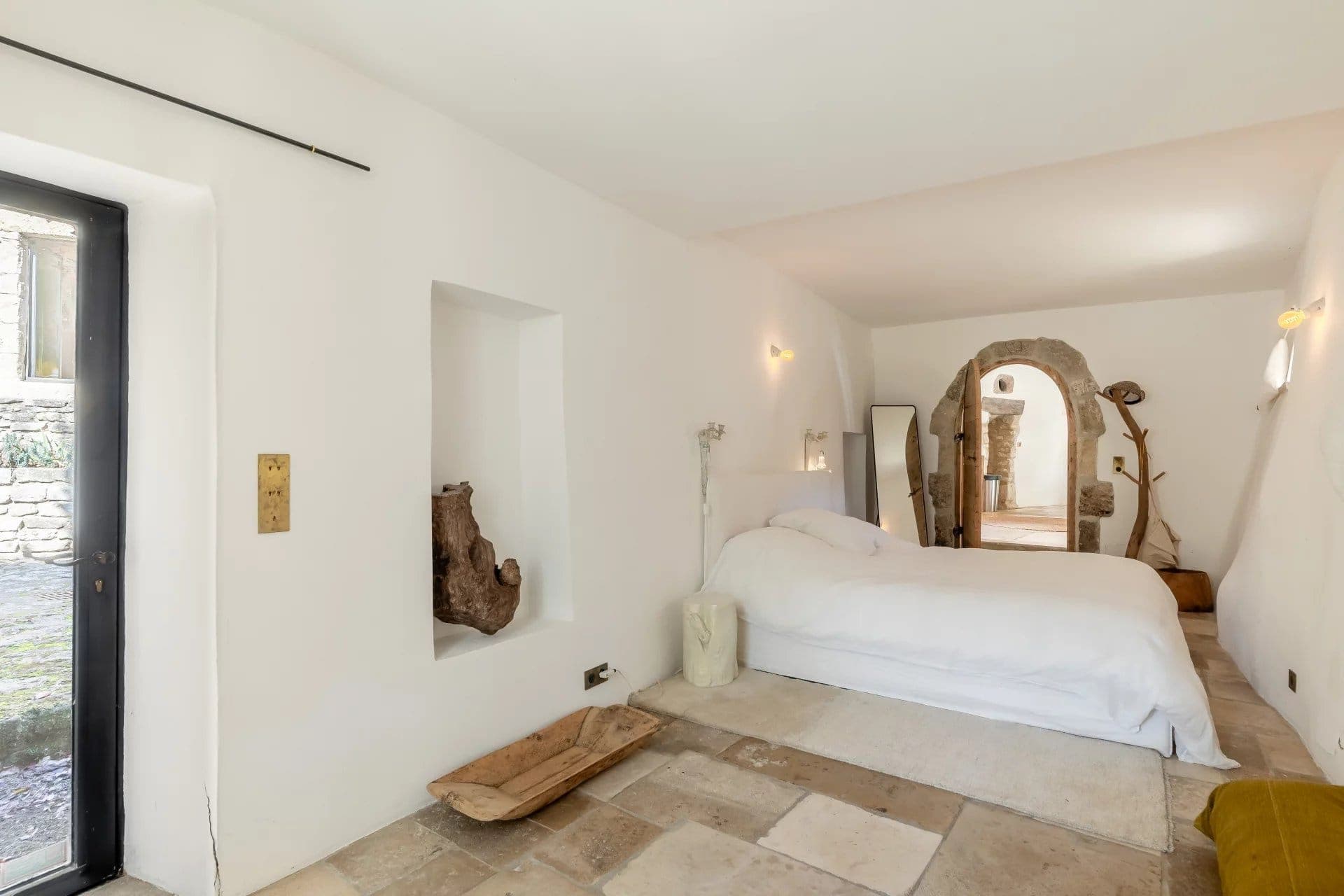 Minimalist bedroom with white walls, featuring a double bed and a stone-tiled floor. There is a large mirror and a rustic wooden bench. An arched doorway leads to another room. Natural light enters through a glass door on the left.