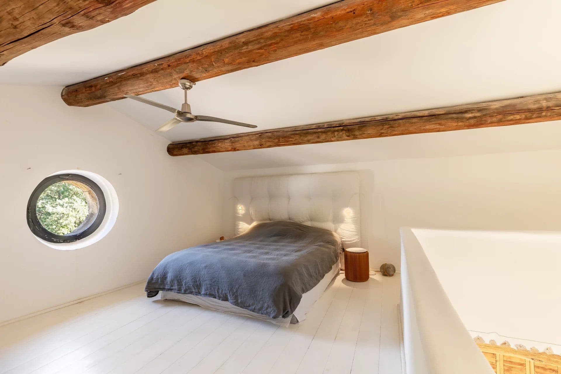 A cozy bedroom with a sloped ceiling featuring exposed wooden beams. A circular window allows natural light in. The bed has a tufted headboard and a gray blanket. A modern ceiling fan hangs overhead, with a small round side table nearby.