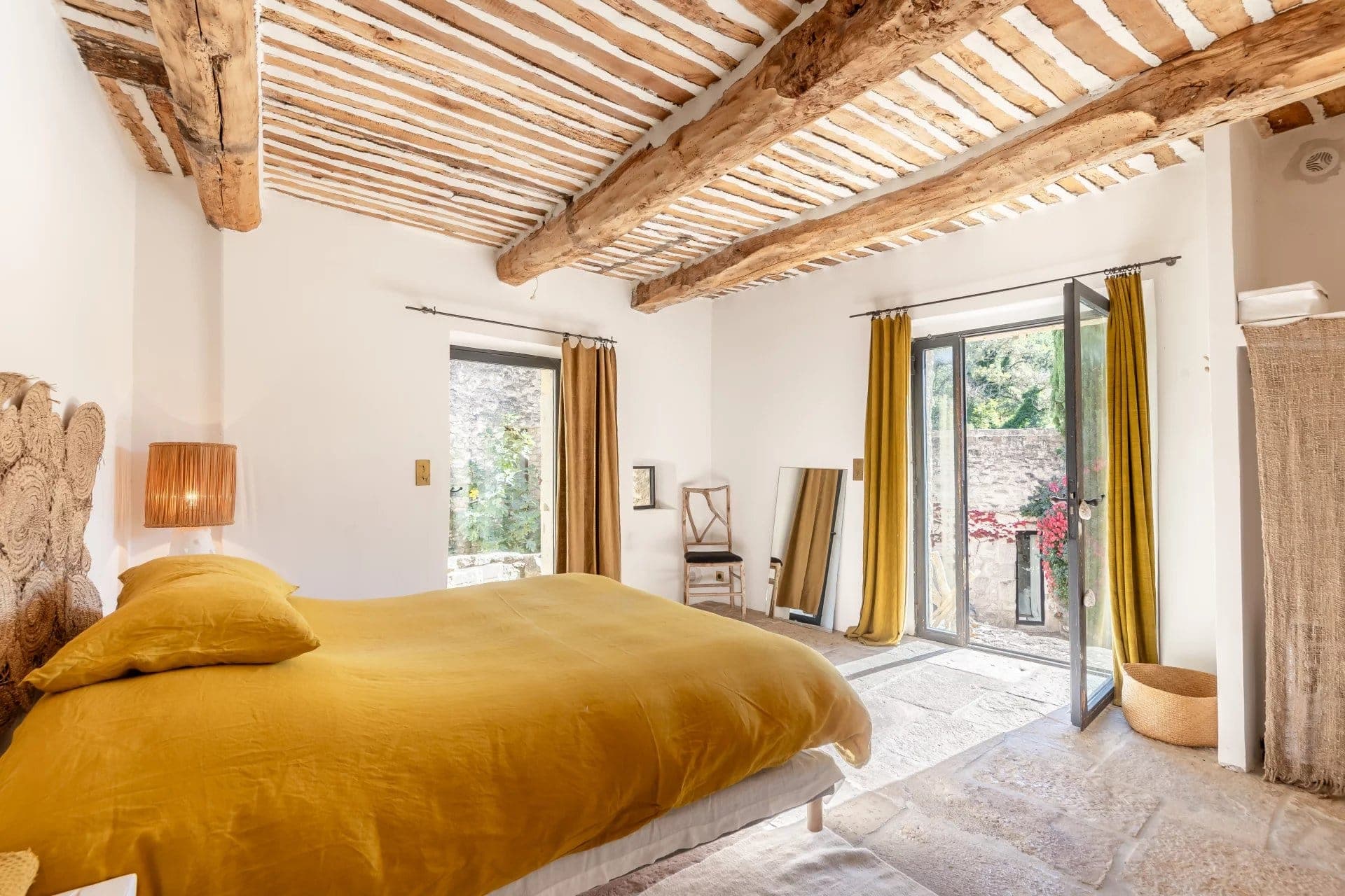 A rustic bedroom features a wooden ceiling with exposed beams, a bed with mustard-yellow bedding, and a standing lamp. A large window in the corner opens to a stone-walled exterior, with natural light illuminating the space.