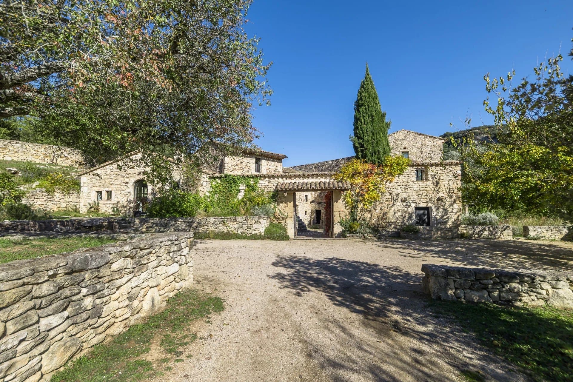 Old stone buildings with arched doorways surrounded by stone walls and trees, set under a clear blue sky. The courtyard and pathways are made of gravel, with greenery adding charm to the rustic setting.