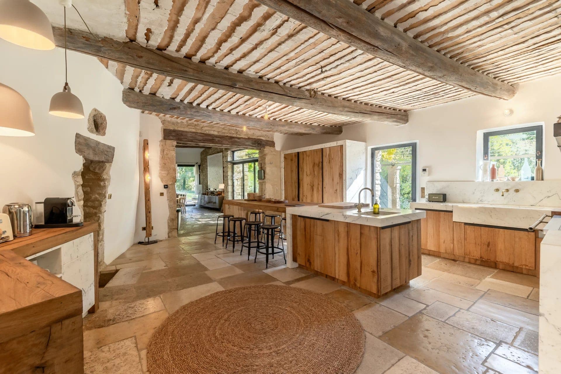 Cuisine spacieuse et rustique avec poutres en bois et sol en pierre naturelle. Îlot central avec tabourets, armoires en bois et grandes fenêtres laissant entrer la lumière naturelle. Les tons neutres et le décor minimaliste créent une atmosphère chaleureuse et accueillante.