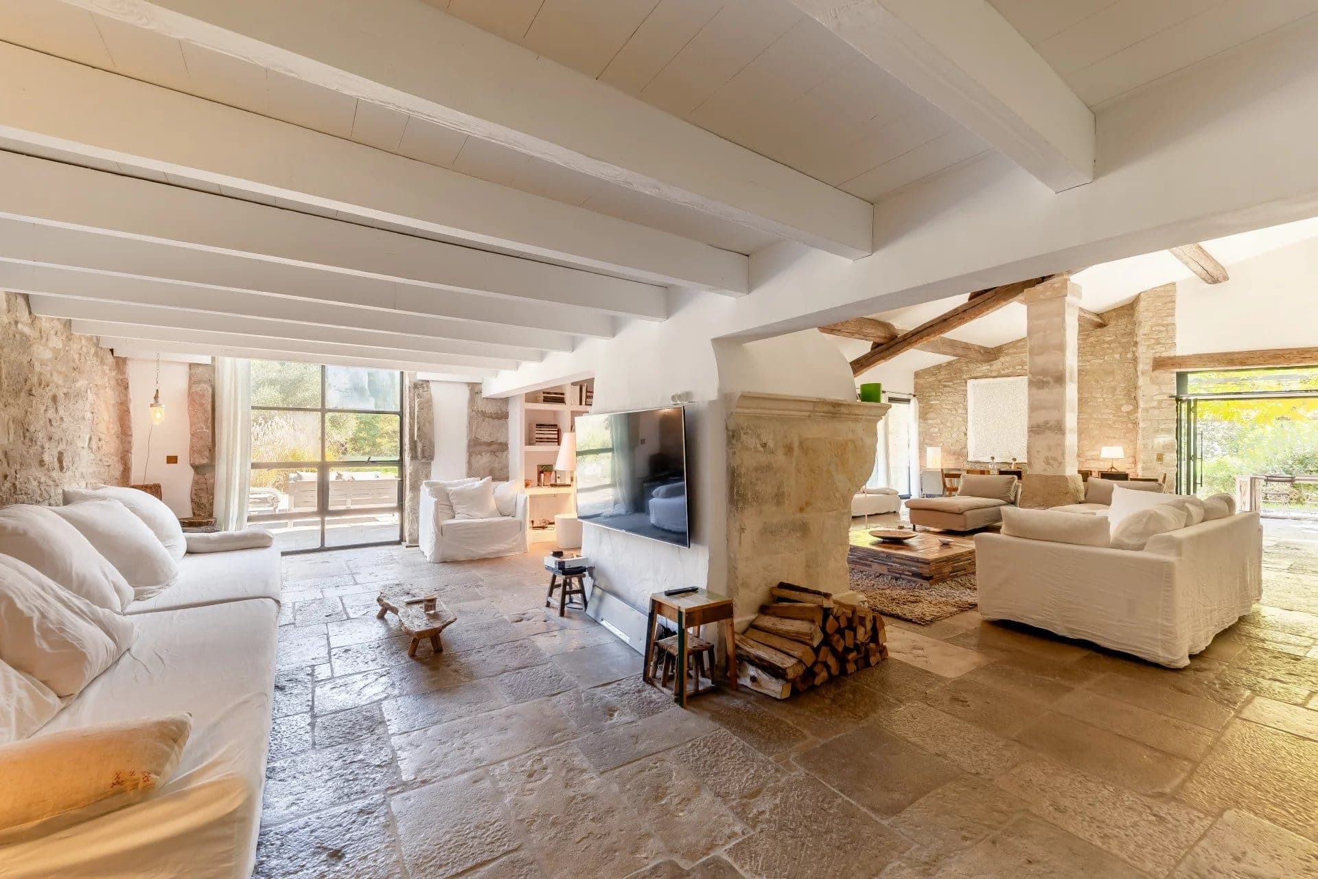 A spacious living room with stone flooring and white walls. It features white sofas, a rustic wooden coffee table, a large TV, and a stacked woodpile. Large windows offer a view of greenery outside, allowing natural light to fill the room.