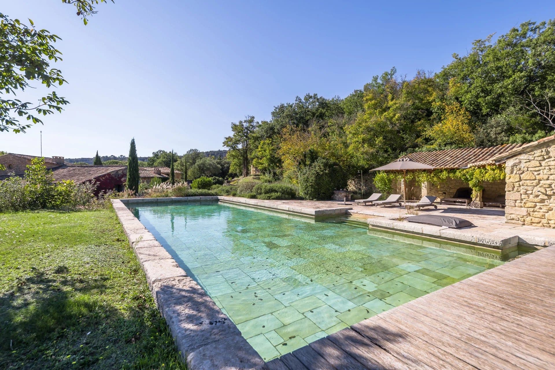 A tranquil outdoor scene features a long, rectangular swimming pool surrounded by a stone deck. Sun loungers sit under a rustic, wooden pergola. Lush greenery and a blue sky complete the serene landscape.