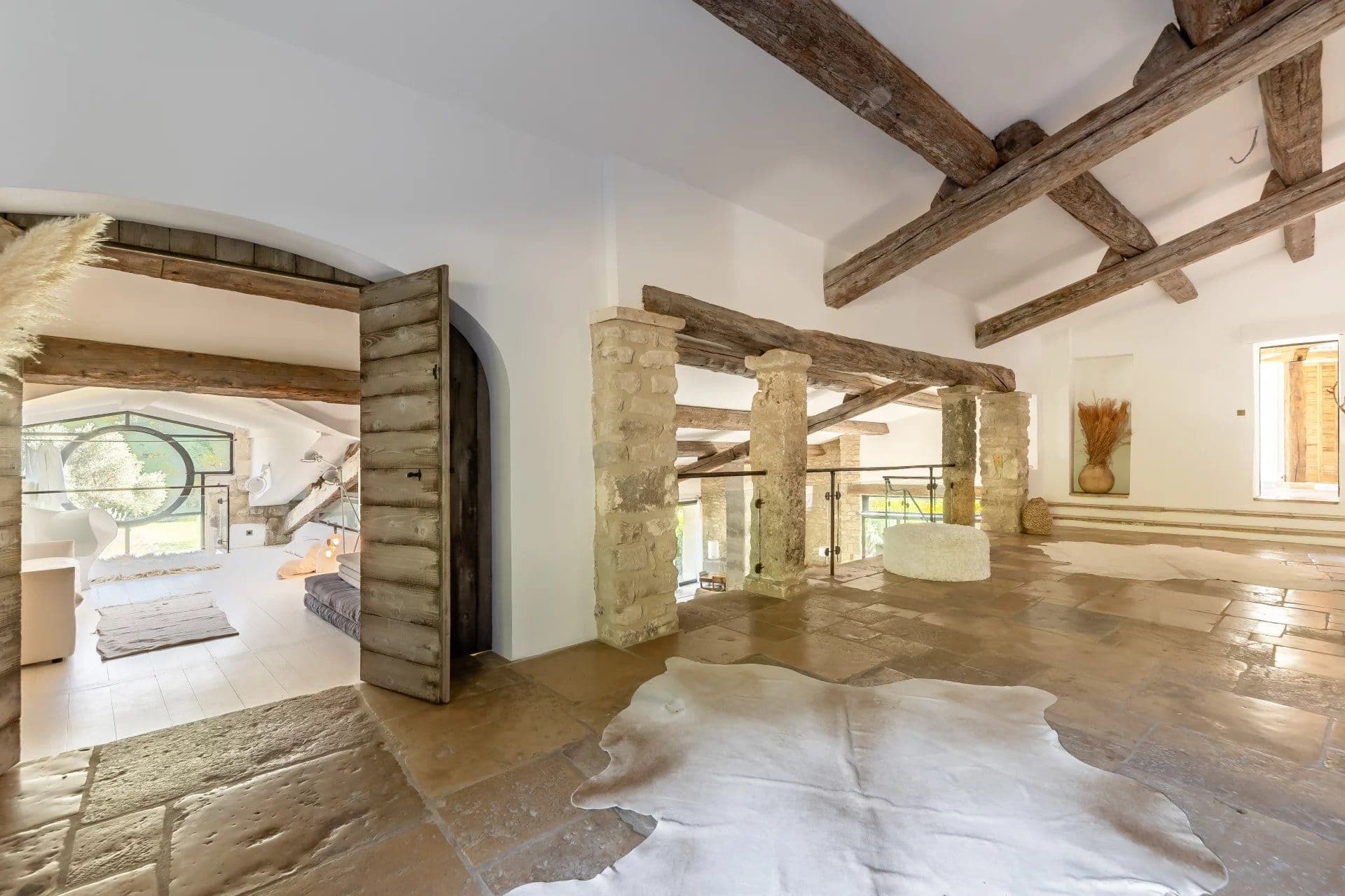 Intérieur spacieux et rustique avec poutres apparentes en bois, piliers en pierre et grandes fenêtres. Un tapis en peau de vache repose sur un sol carrelé. La lumière naturelle remplit l'espace, mettant en valeur un mélange d'éléments de décoration modernes et traditionnels.