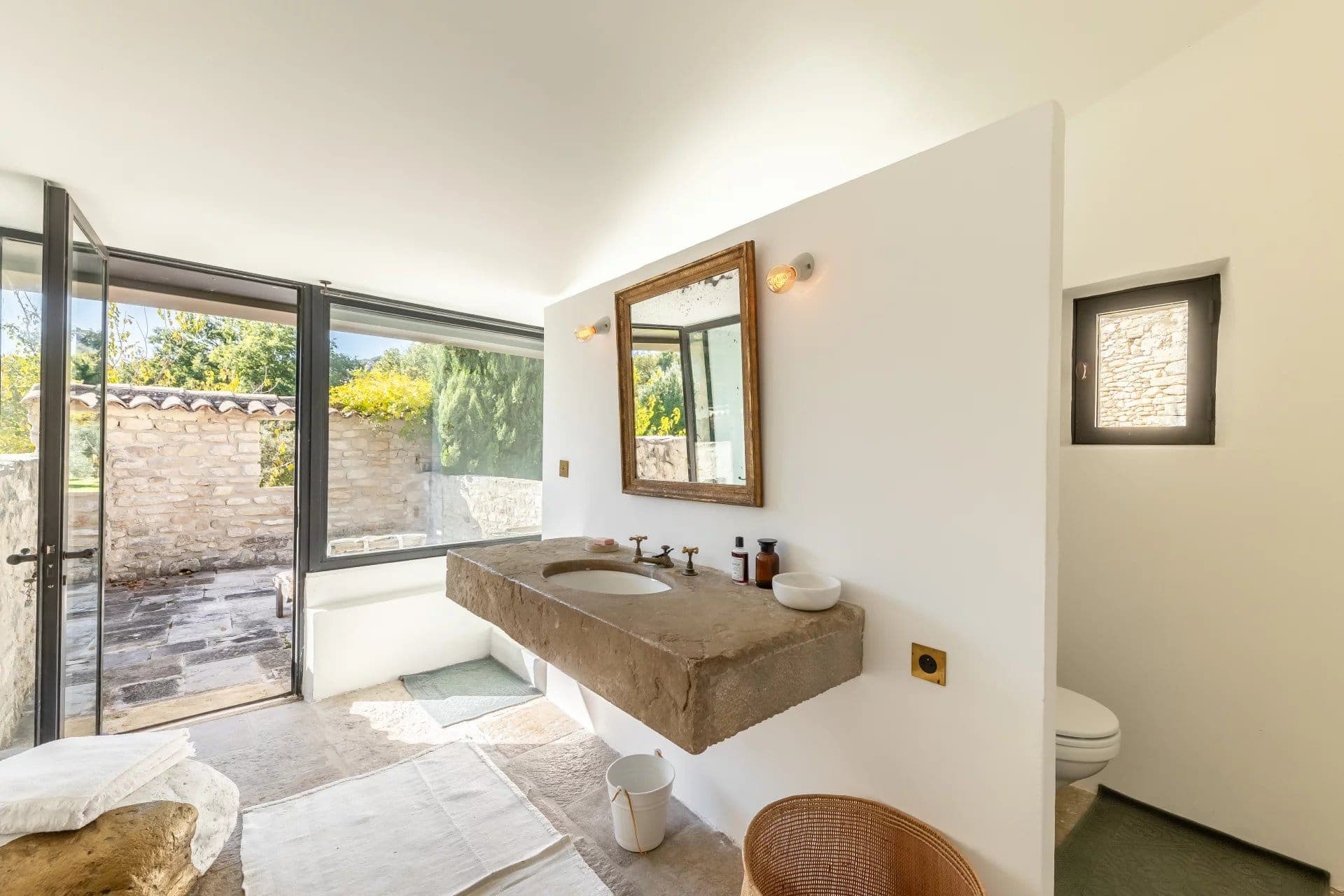 Bright bathroom with a large stone sink and countertop, wooden mirror, and modern fixtures. Sunlight streams through glass doors leading to a stone-paved patio with greenery. A white rug and wicker basket add warmth to the stone floor.