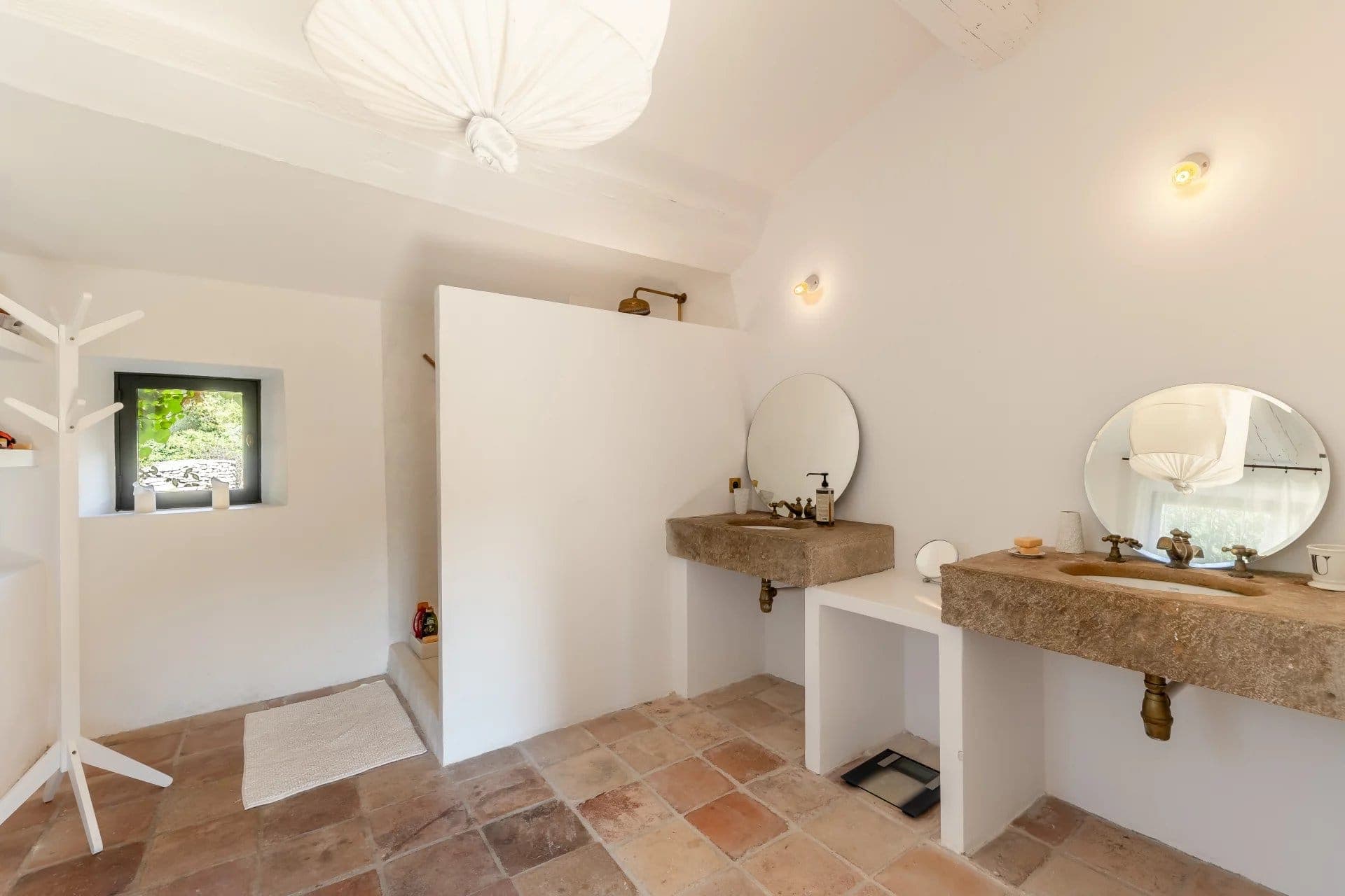 A minimalist bathroom features two stone sinks with round mirrors, a walk-in shower with a partial wall, and terracotta floor tiles. Soft lighting and a hanging fabric lamp create a serene atmosphere. A window provides natural light and greenery views.
