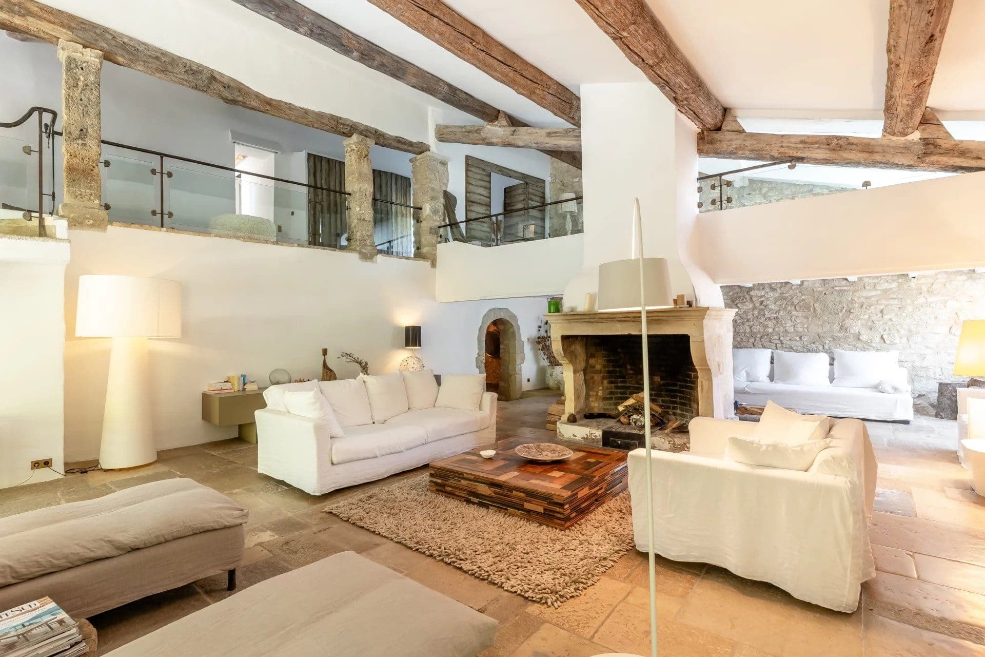 A spacious living room with rustic charm, featuring exposed wooden beams and stone walls. The room has cozy white sofas, a large fireplace, wooden coffee tables, and soft lighting, creating a warm and inviting atmosphere.