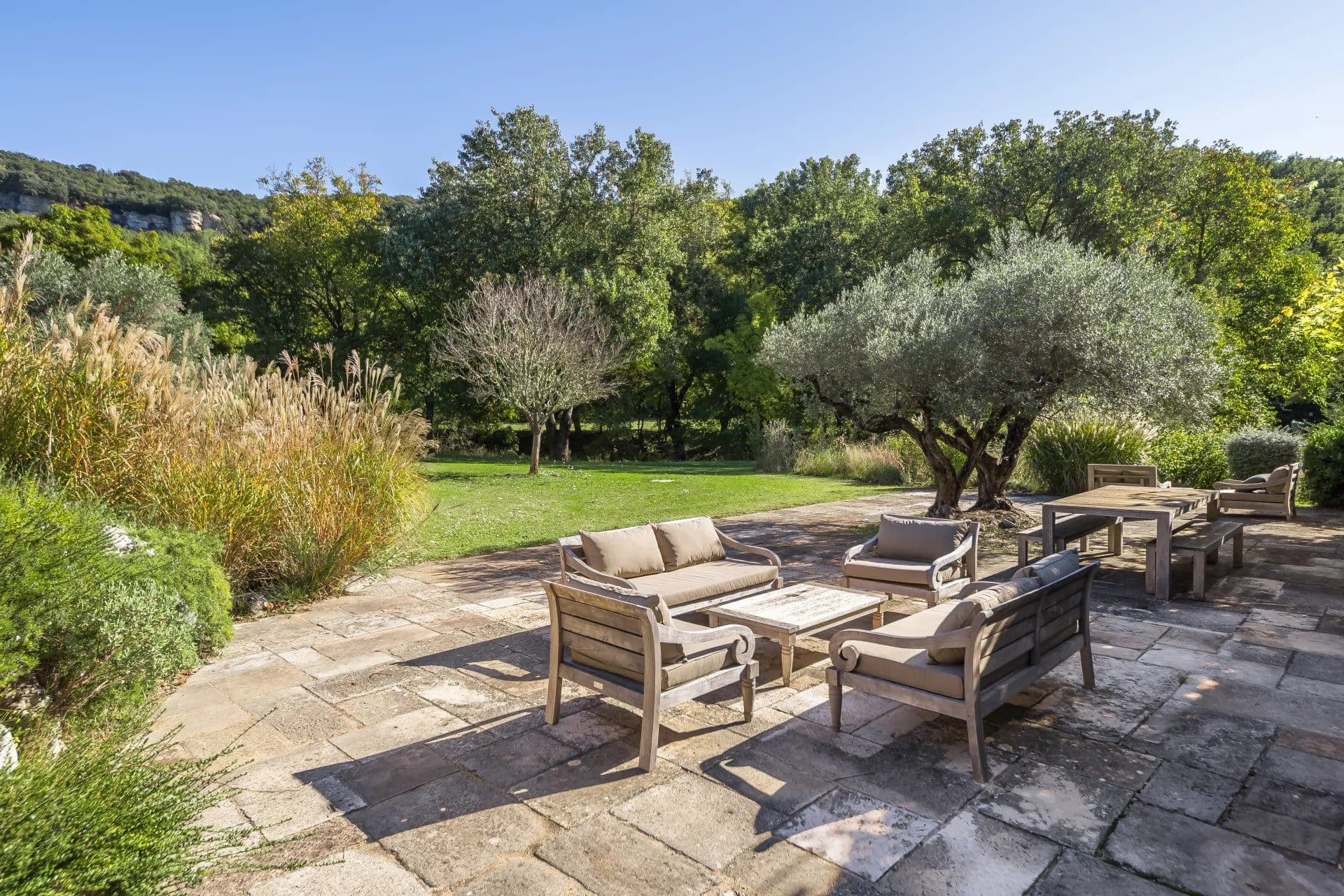 A rustic outdoor patio with cushioned wooden seating surrounds a wooden coffee table. The patio is set on stone tiles, overlooking a lush green lawn with trees and bushes under a clear blue sky.