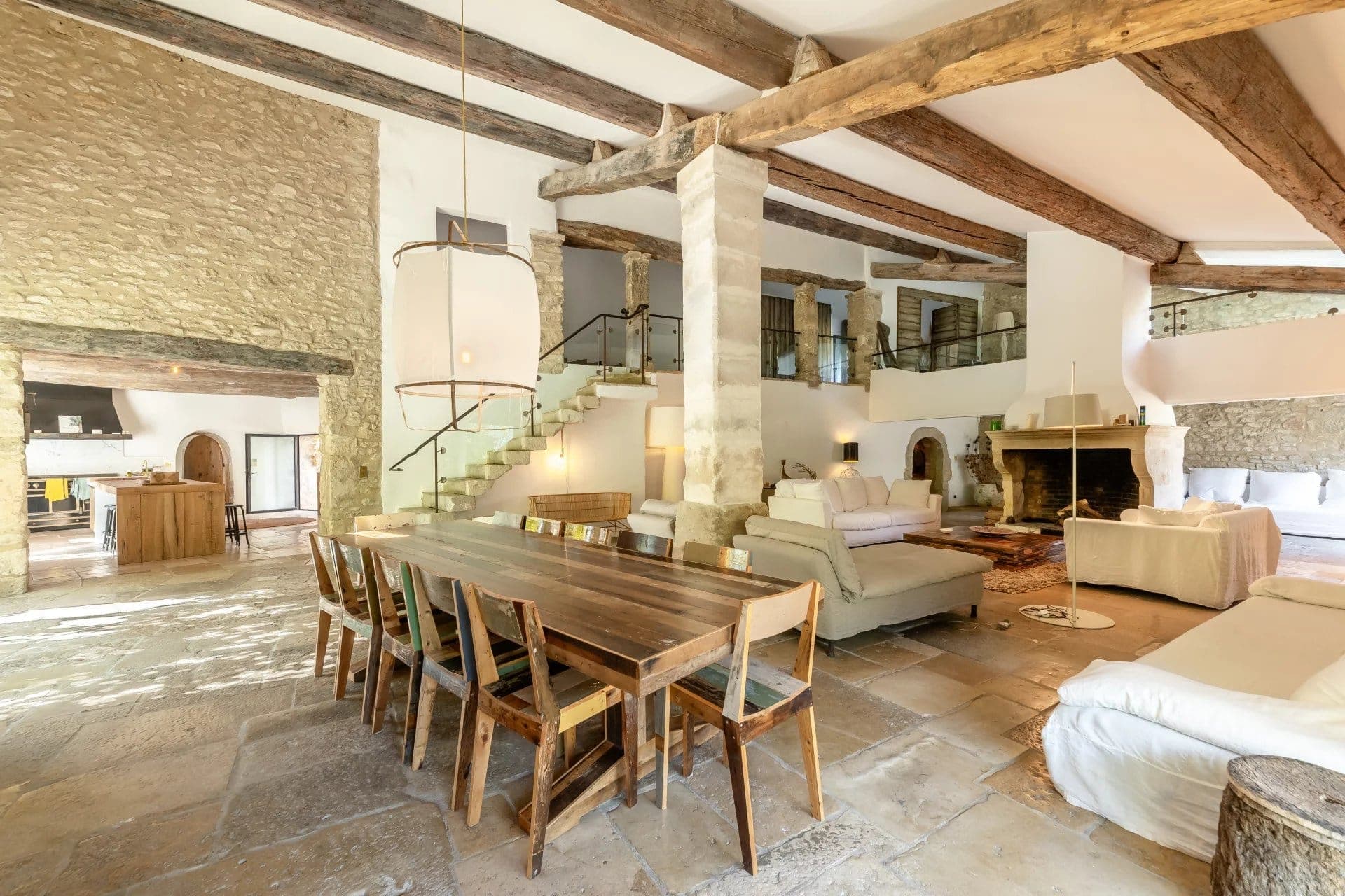 Salon et salle à manger rustiques avec murs en pierre et poutres en bois. Une longue table en bois avec des chaises se trouve à gauche et des canapés blancs près d'une cheminée se trouvent à droite. Un escalier mène à un niveau supérieur. La lumière naturelle illumine l'espace.